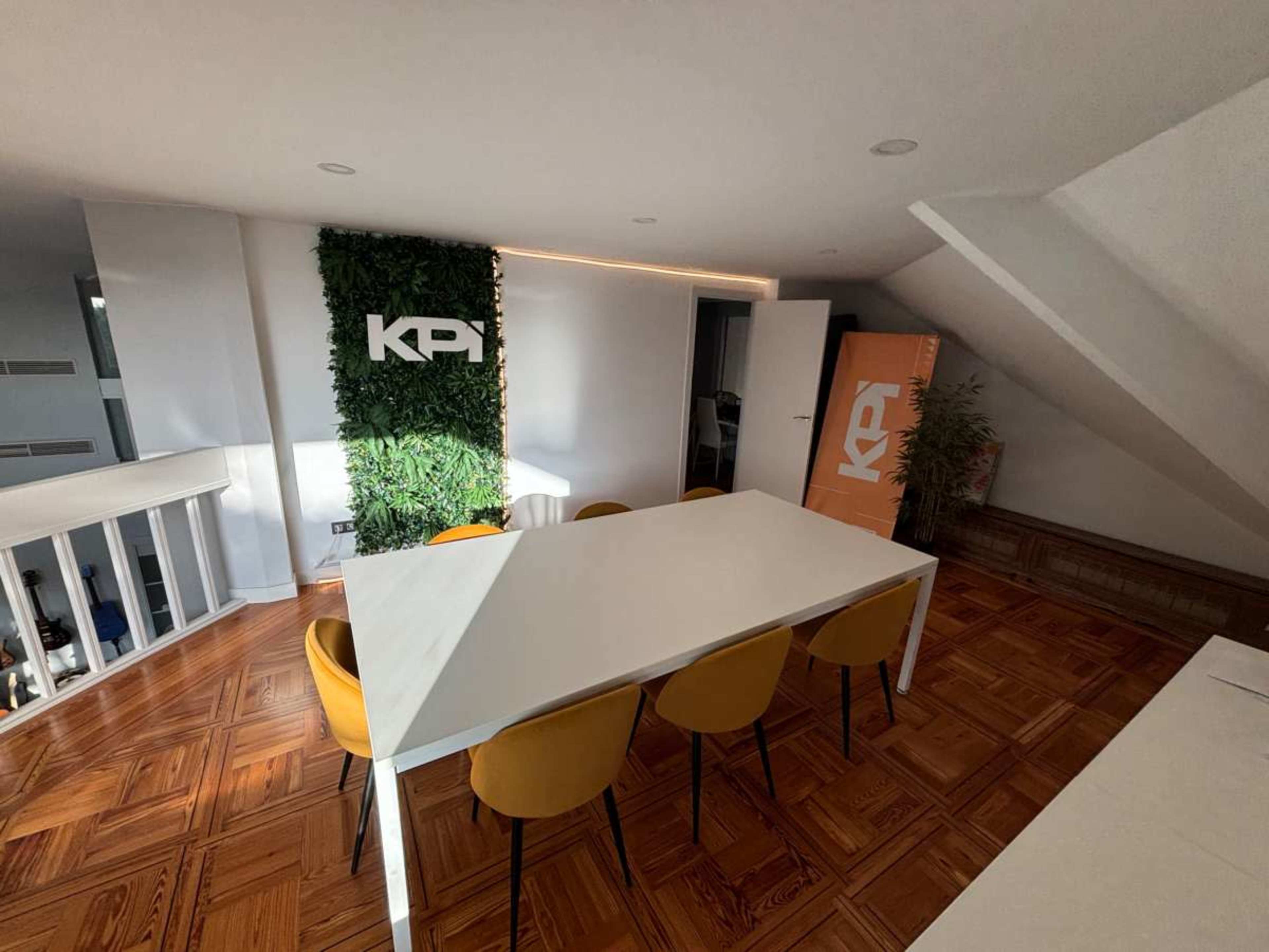 The image shows a modern meeting room with a large white table surrounded by yellow chairs, featuring a green wall with the letters "KPI" and an orange banner in the corner.
