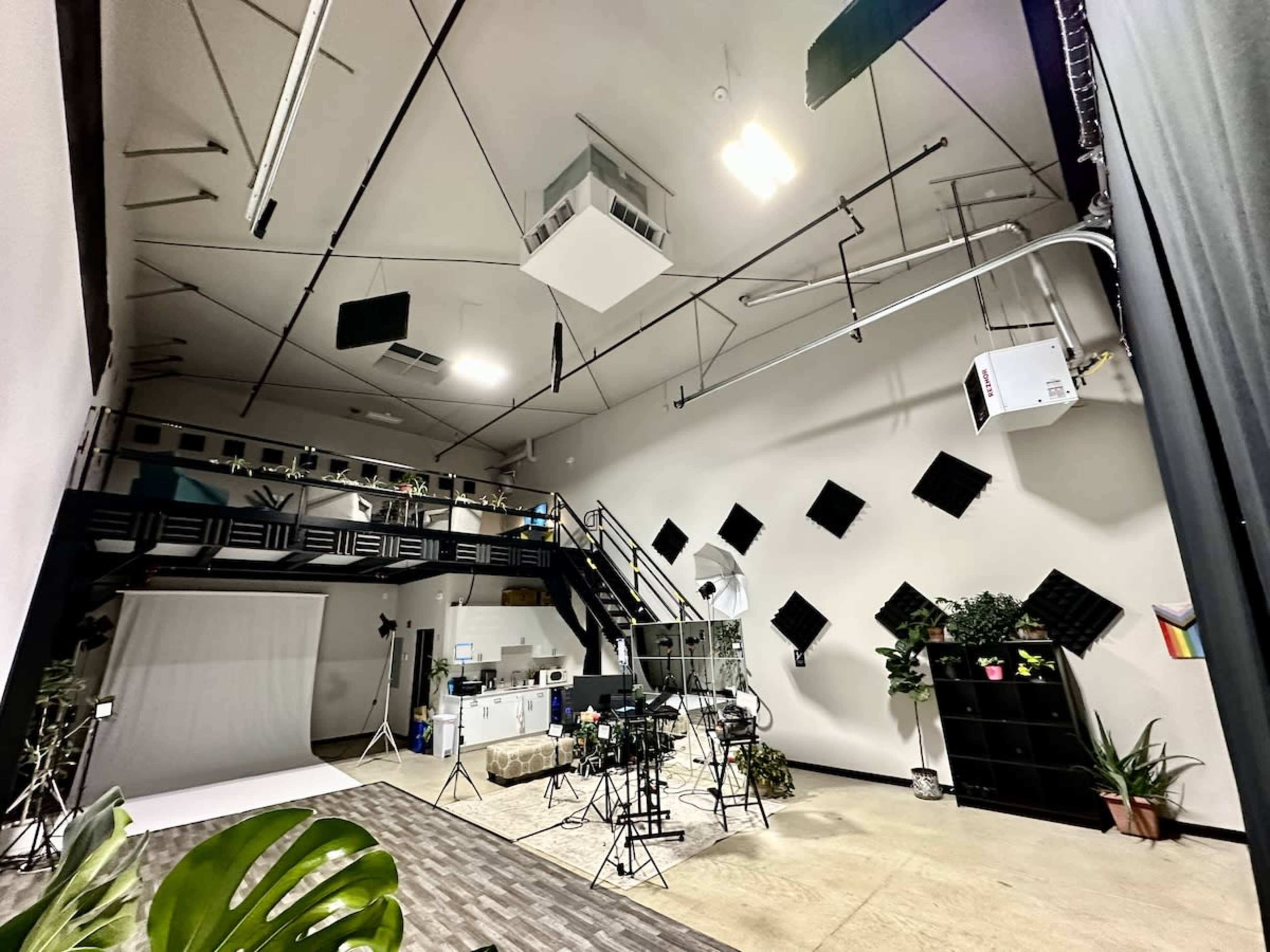 The image shows a spacious studio with a multi-level setup, featuring various lighting and filming equipment, a backdrop area, and plants arranged throughout the space.