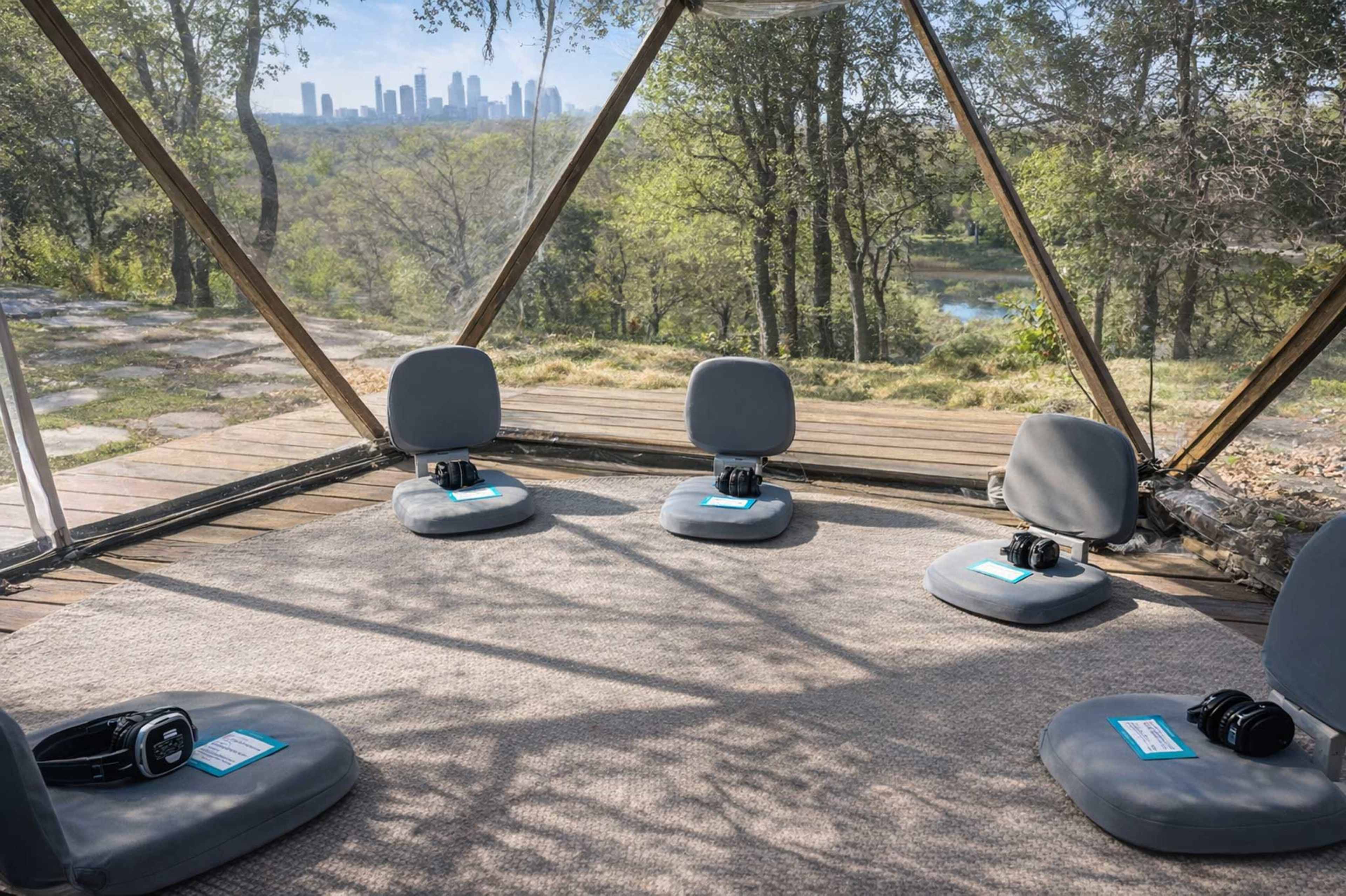The image shows a transparent geodesic dome with five cushioned seats arranged on a carpet, overlooking a wooded area and a skyline in the distance.