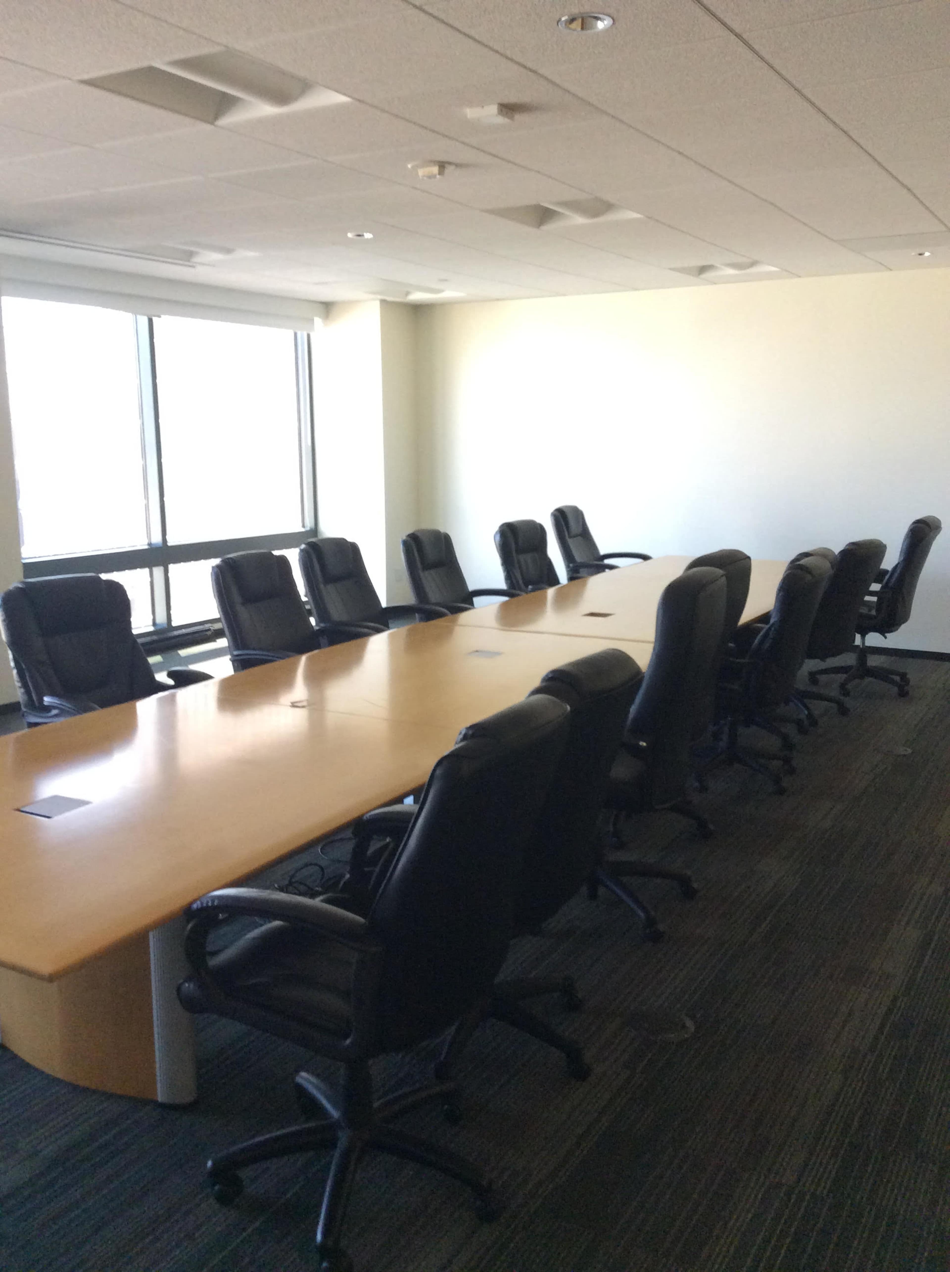 A large conference room features a long wooden table surrounded by black ergonomic chairs and a large window providing natural light.