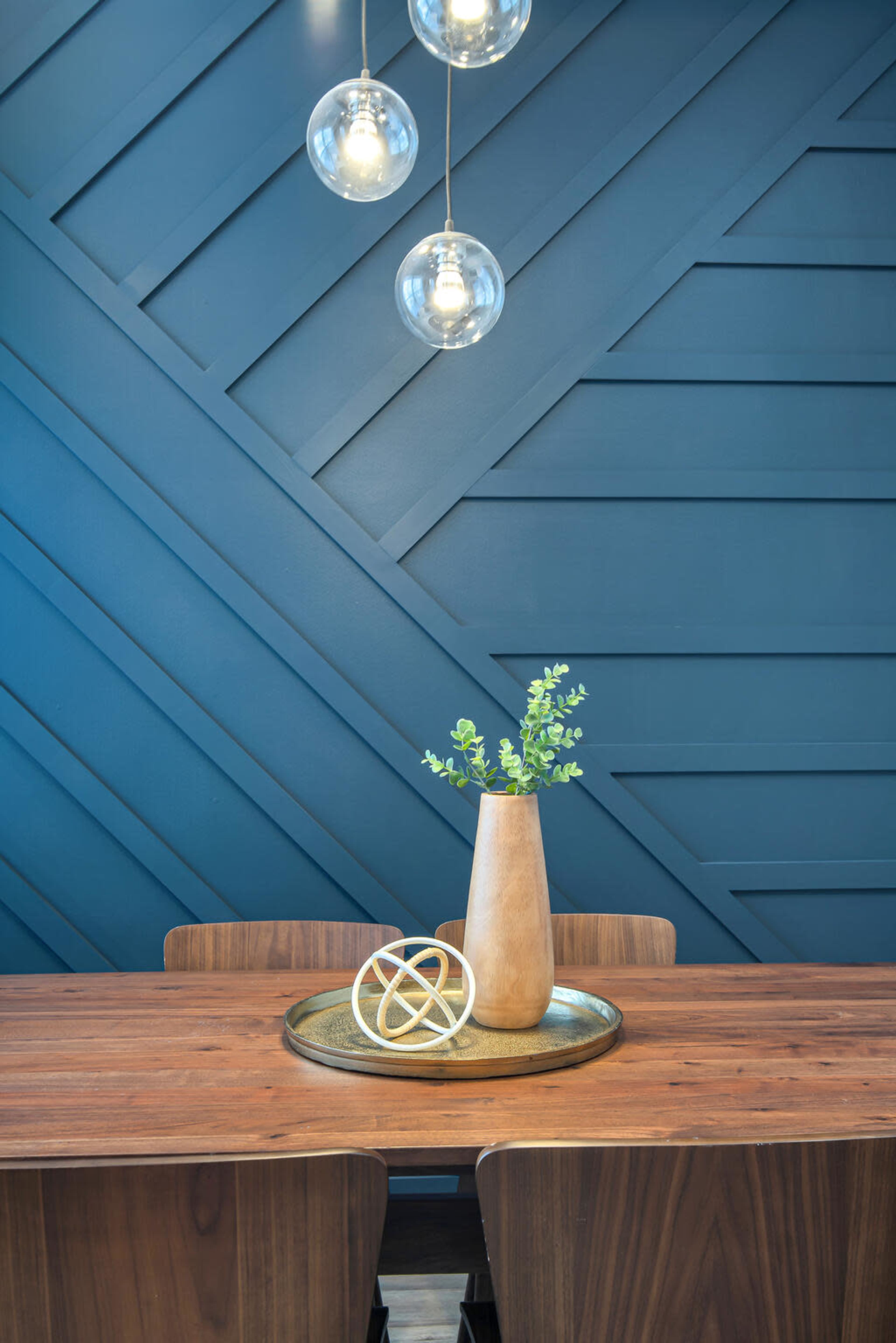 A wooden table is set with a round tray holding a decorative item and a vase containing green foliage, against a blue geometric-patterned wall illuminated by pendant lights.