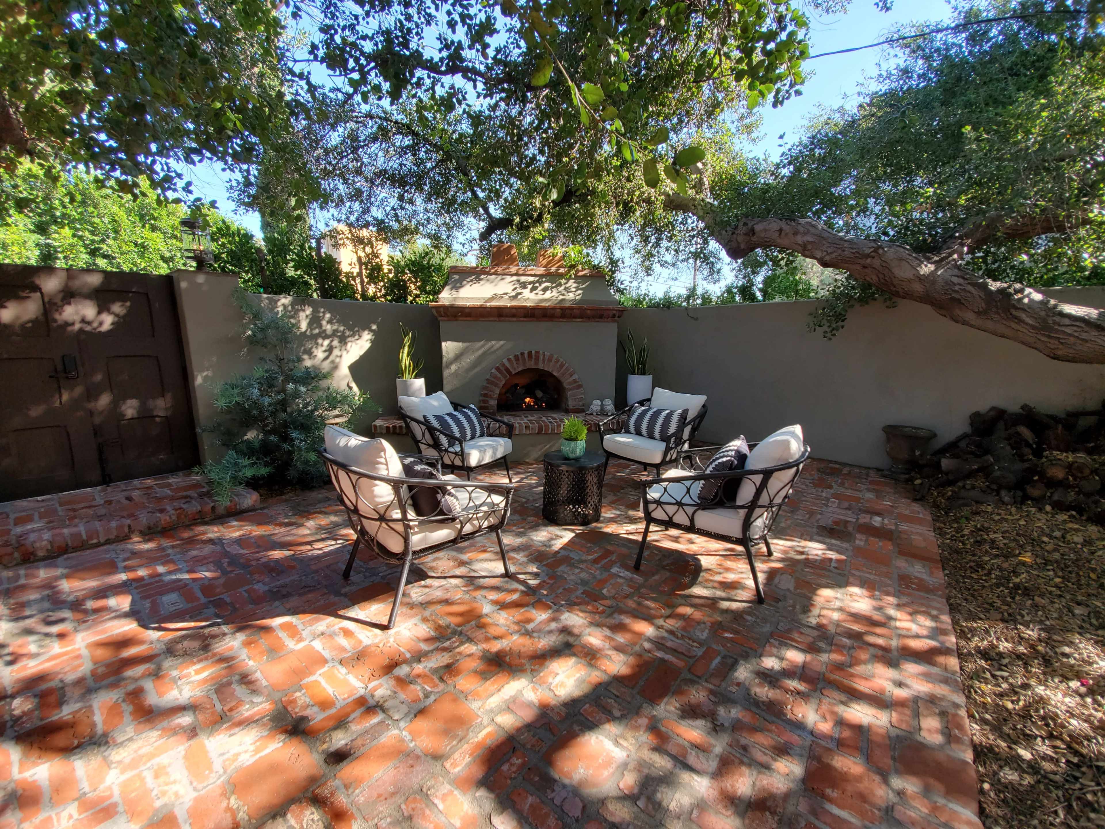 The image shows a patio area with four chairs arranged around a table, a brick floor, and a fireplace surrounded by greenery.