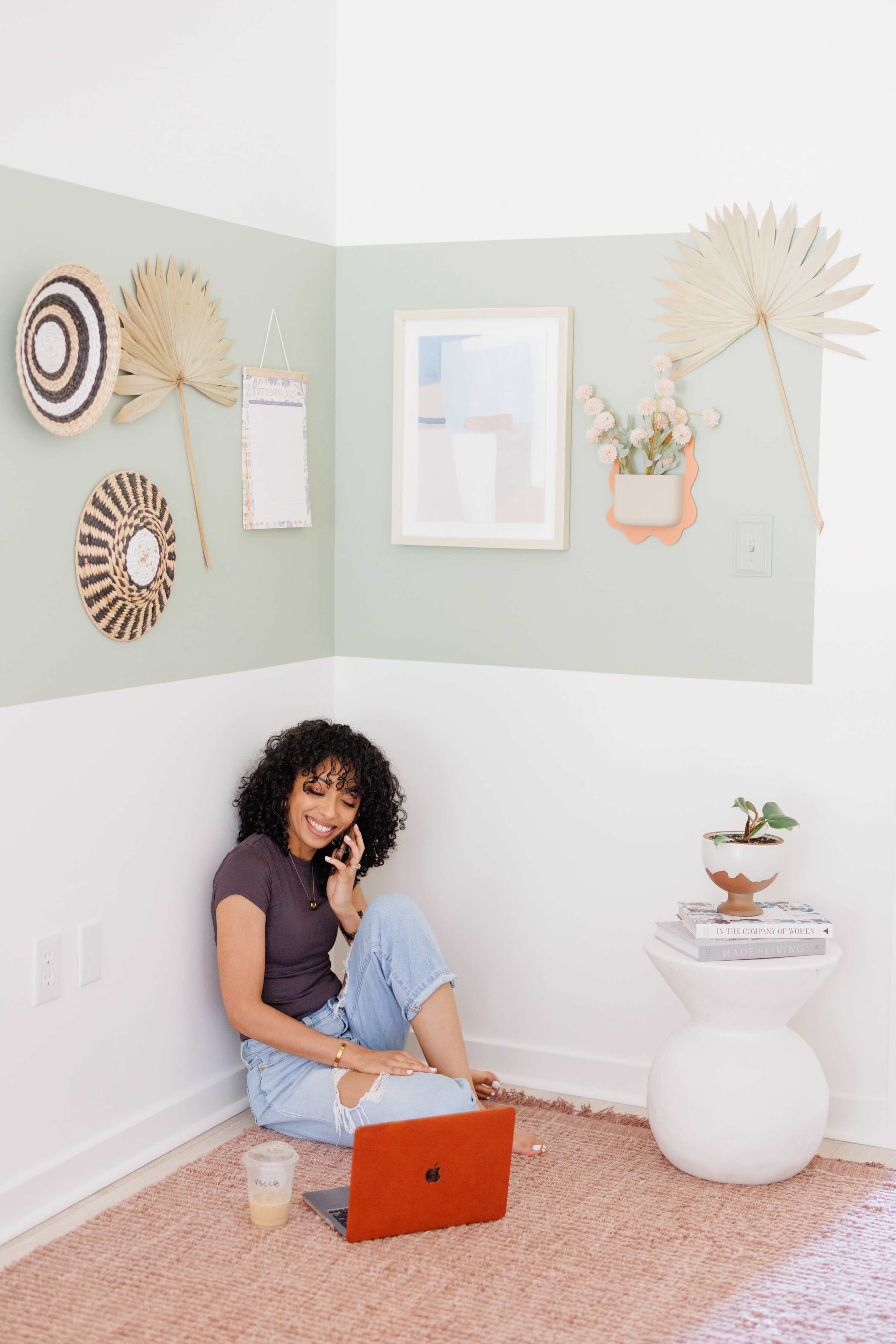 A person sits in a corner of a room with a laptop, surrounded by decorative wall art and a small potted plant.