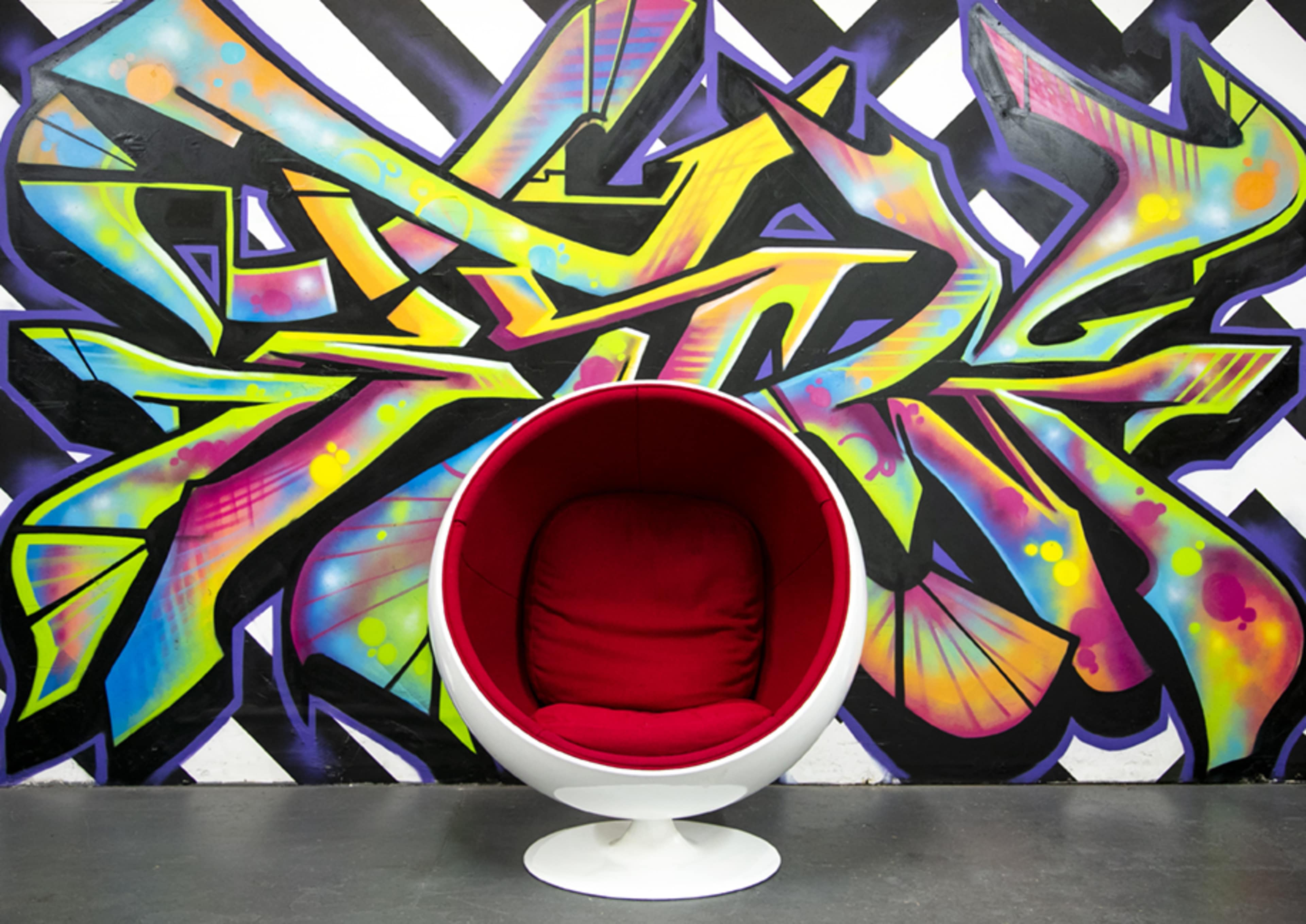 A round red chair is positioned in front of a vibrant, multicolored graffiti wall featuring angular designs.