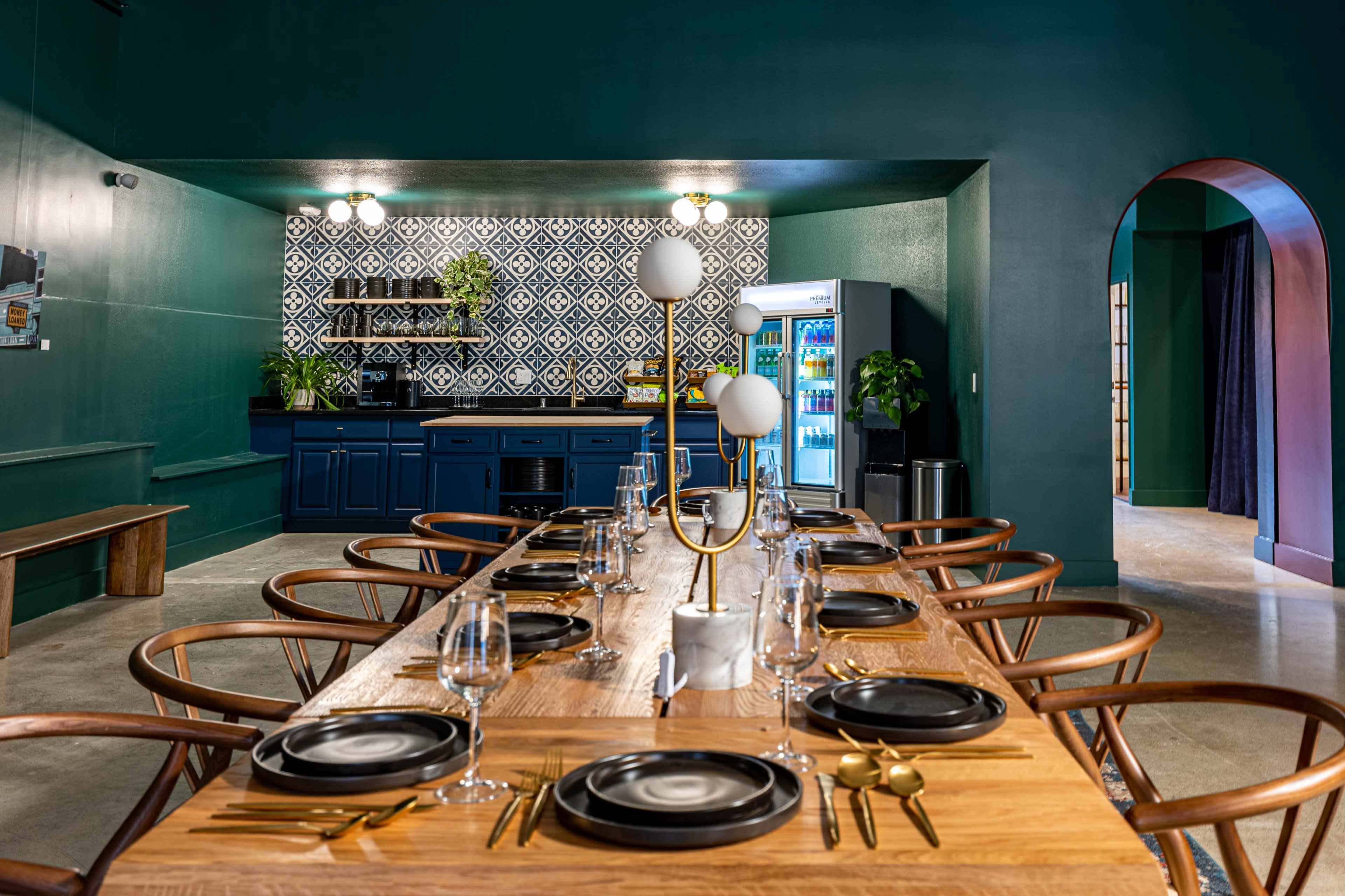 A modern dining area features a long wooden table set with plates and glassware, surrounded by stylish chairs, with a blue kitchen area and patterned backsplash in the background.
