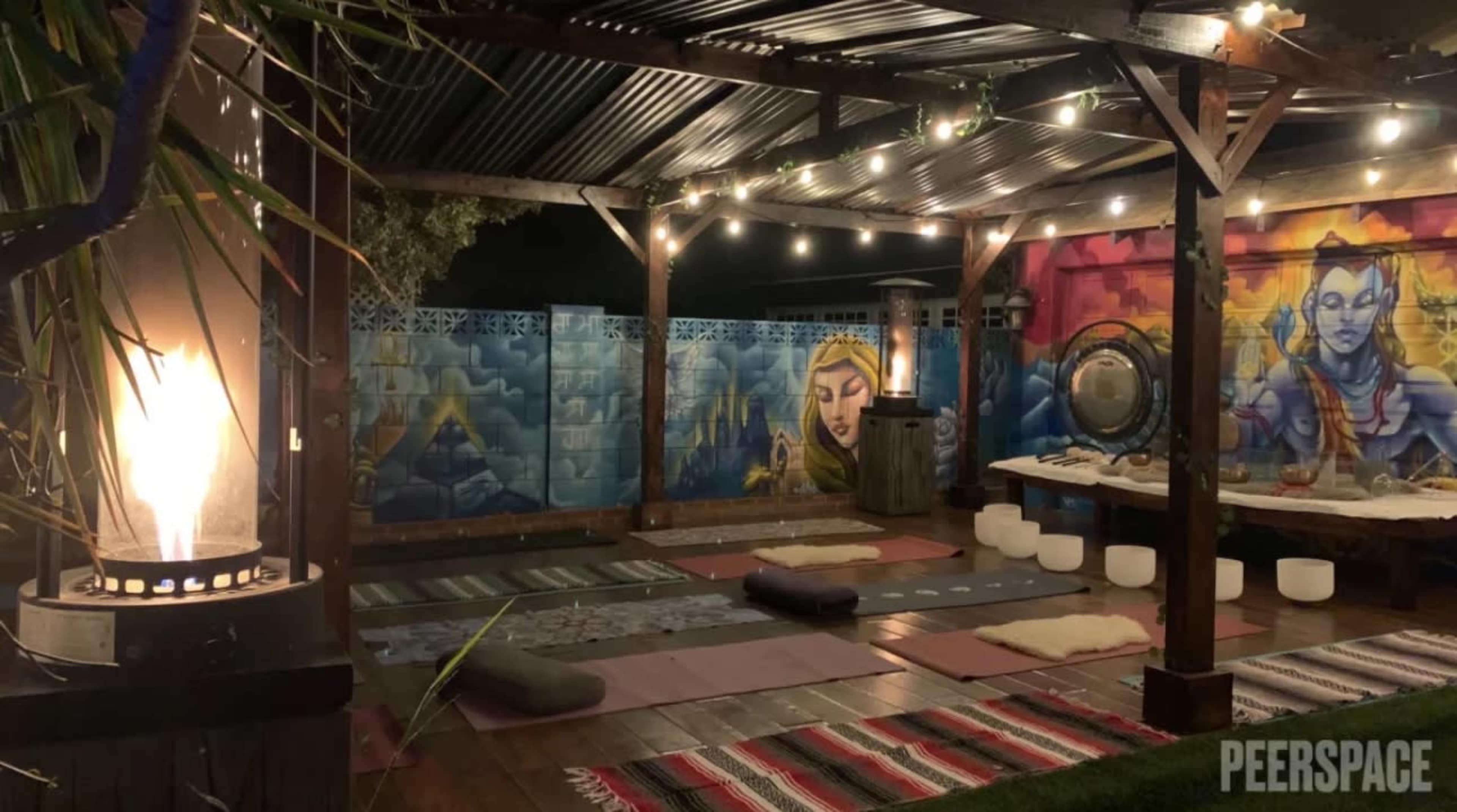 The outdoor space features a decorative wooden structure with colorful murals, seating mats, and a fire feature, all illuminated by string lights.