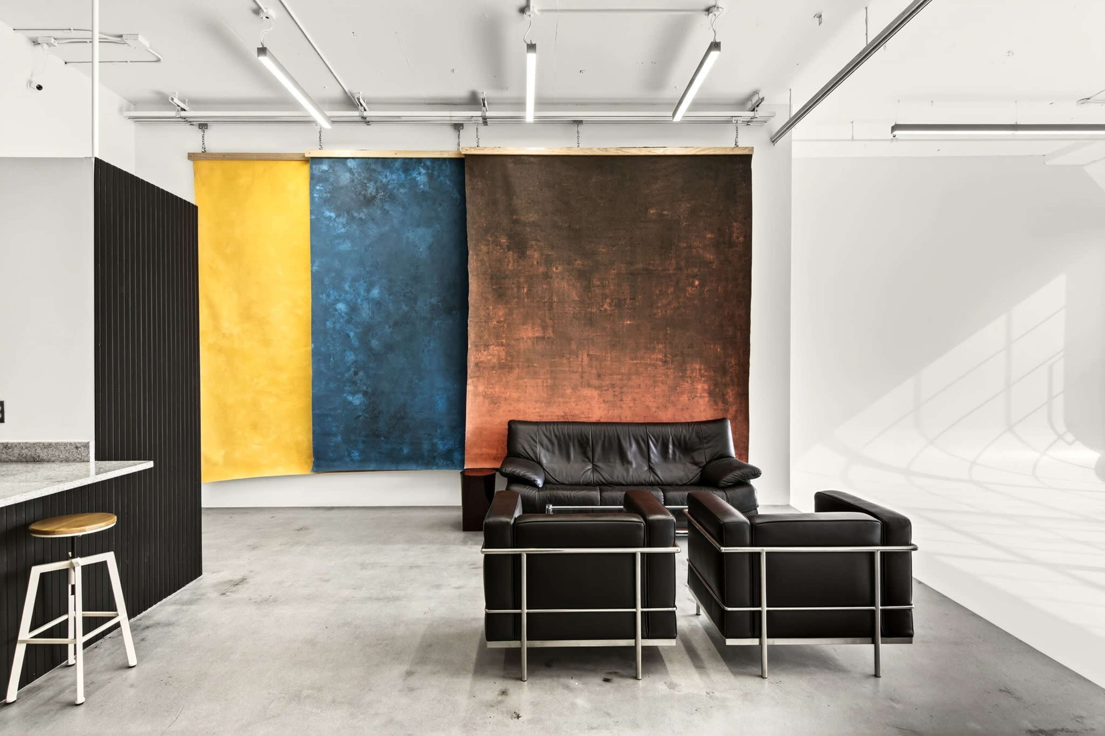 The image shows a modern interior space with a black leather sofa and armchair, complemented by large abstract fabric backdrops in blue, yellow, and brown.