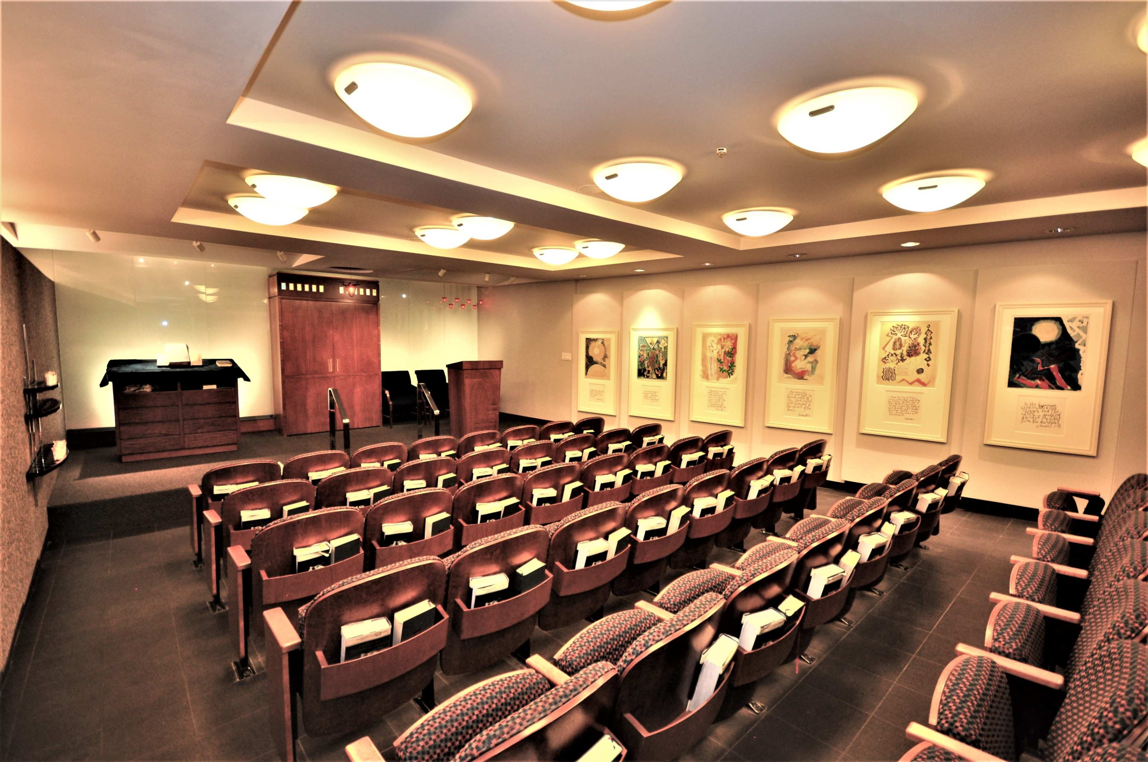 The image shows a conference room with rows of wooden chairs facing a podium, flanked by framed artworks on the walls.