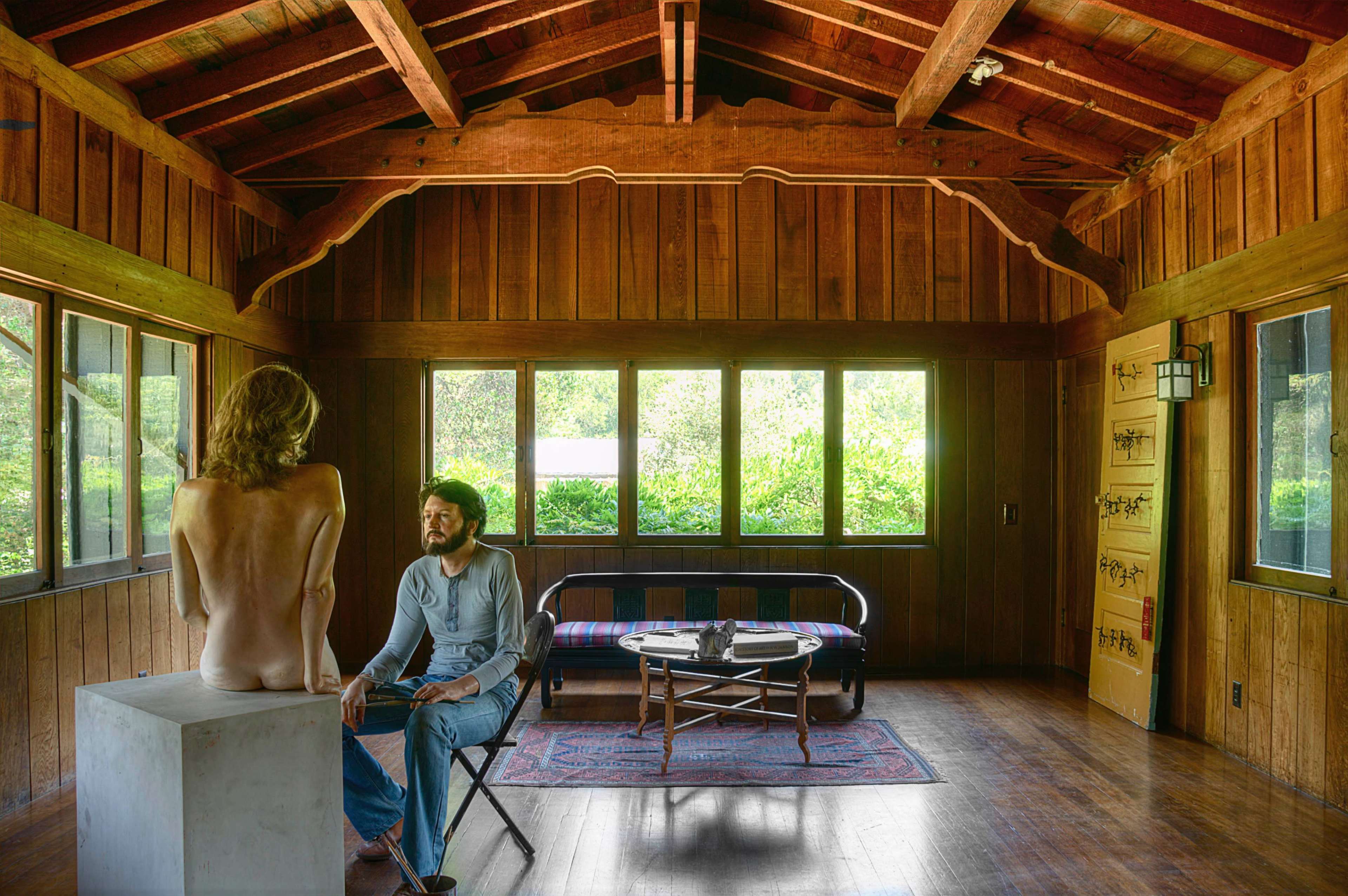 A person is seated on a chair facing a nude model perched on a pedestal in a wooden cabin with large windows overlooking greenery.