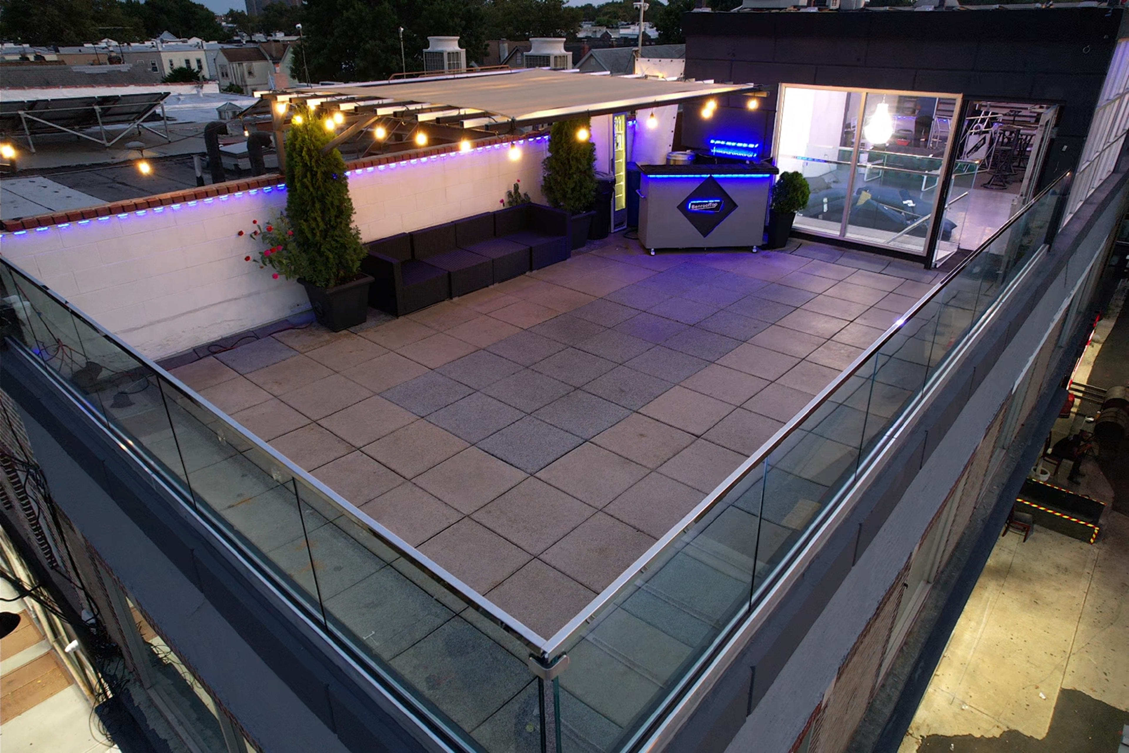 A rooftop terrace featuring a lounge area with seating, plants, and a lighted counter, surrounded by glass railings.