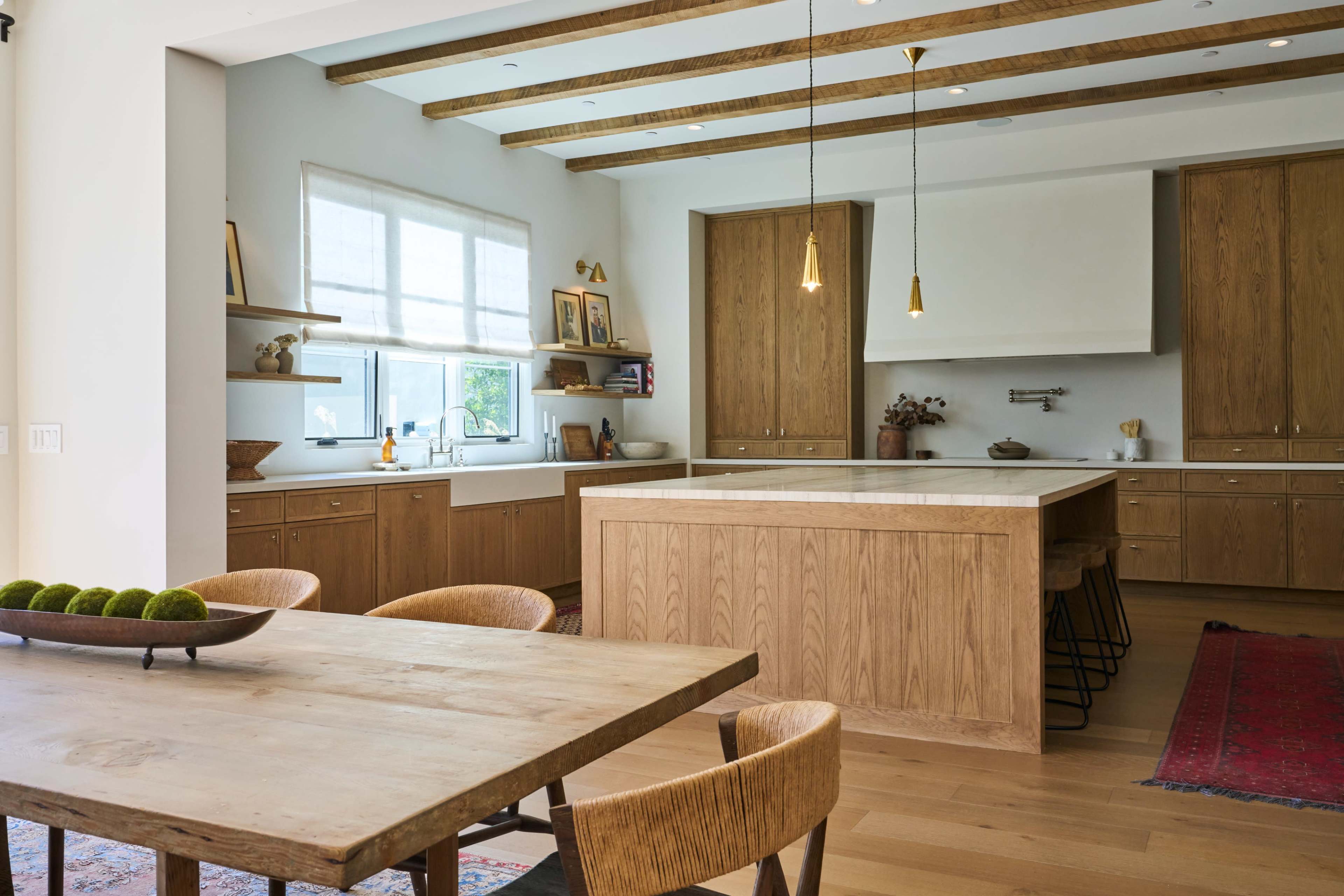 The image shows a modern kitchen and dining area with wooden cabinetry, a large island, and a wooden dining table surrounded by chairs.