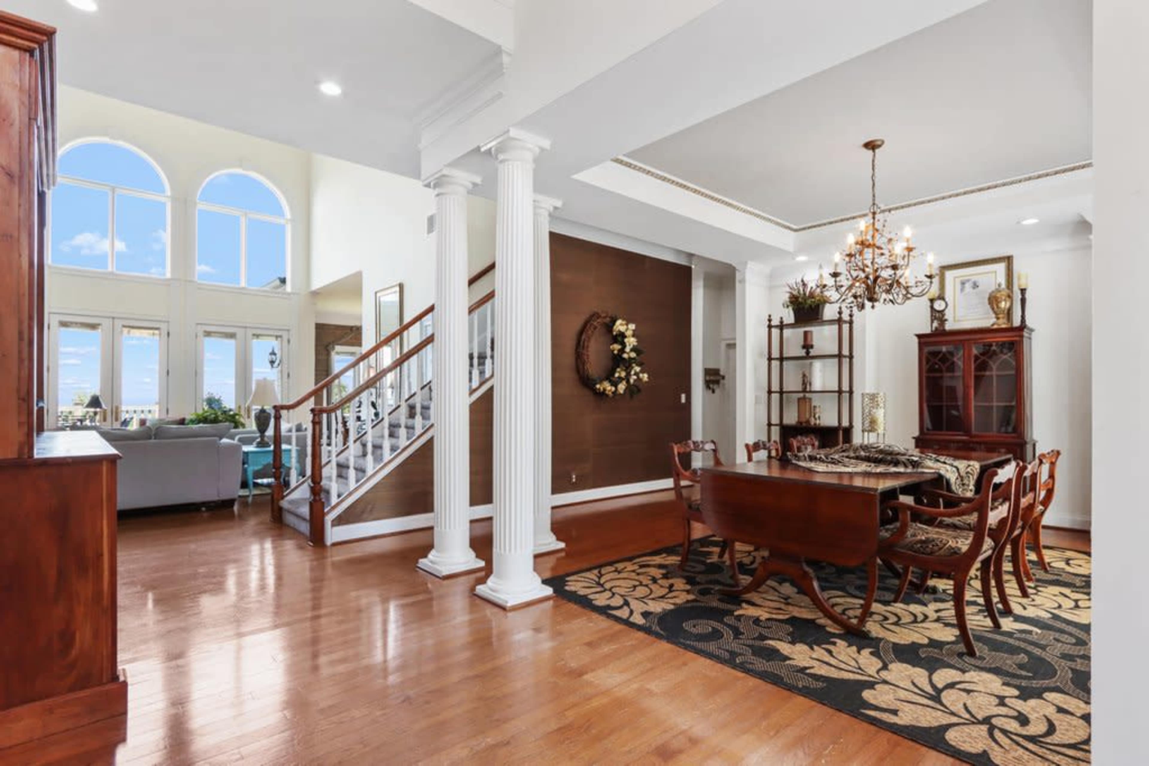 An elegant dining area with a wooden table and chairs, adjacent to a staircase and large windows allowing natural light to fill the space.