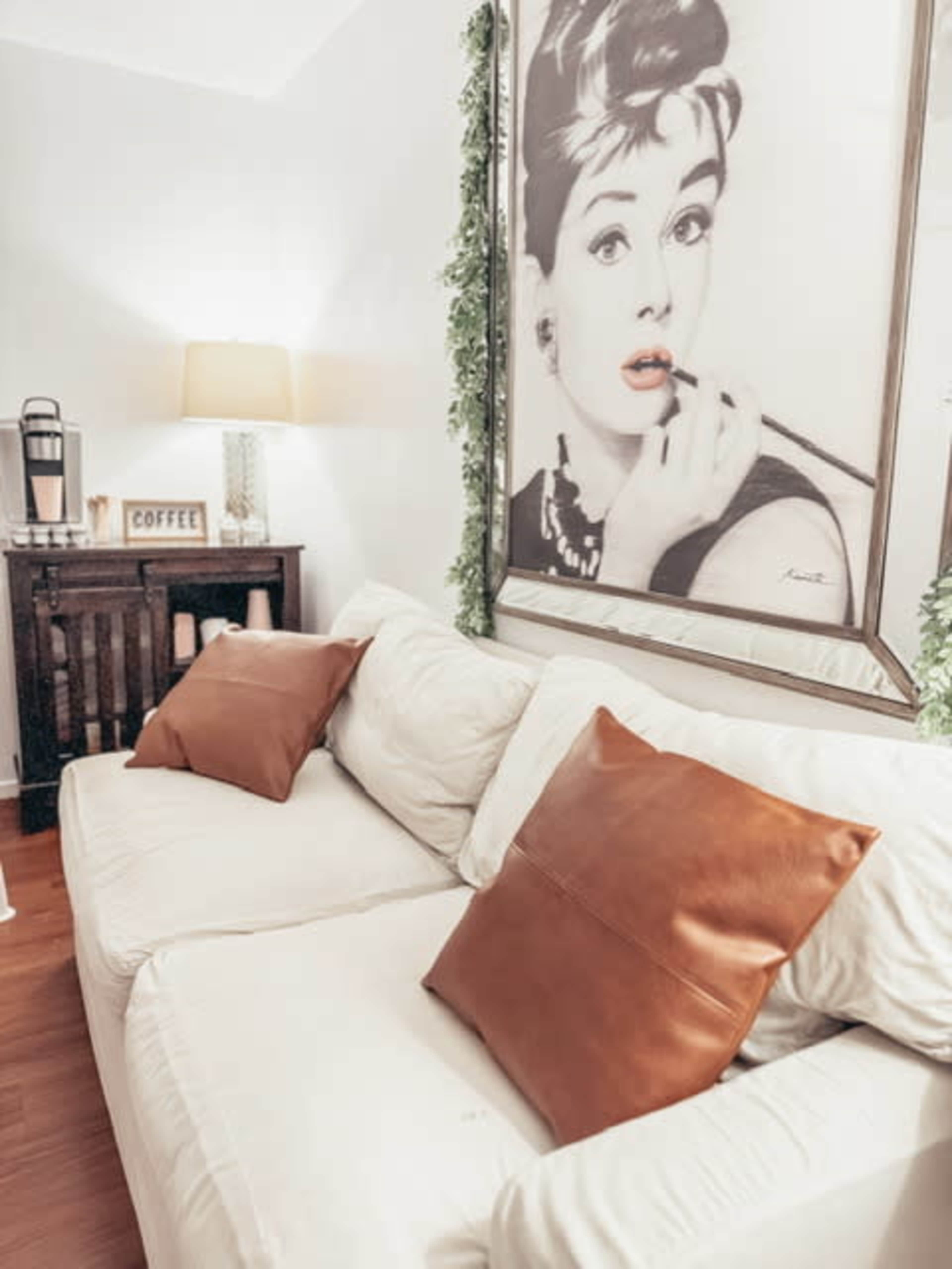 A white sofa with two brown pillows sits against a wall featuring a large framed image of a woman holding a cigarette holder, alongside a lamp and a coffee station in the background.