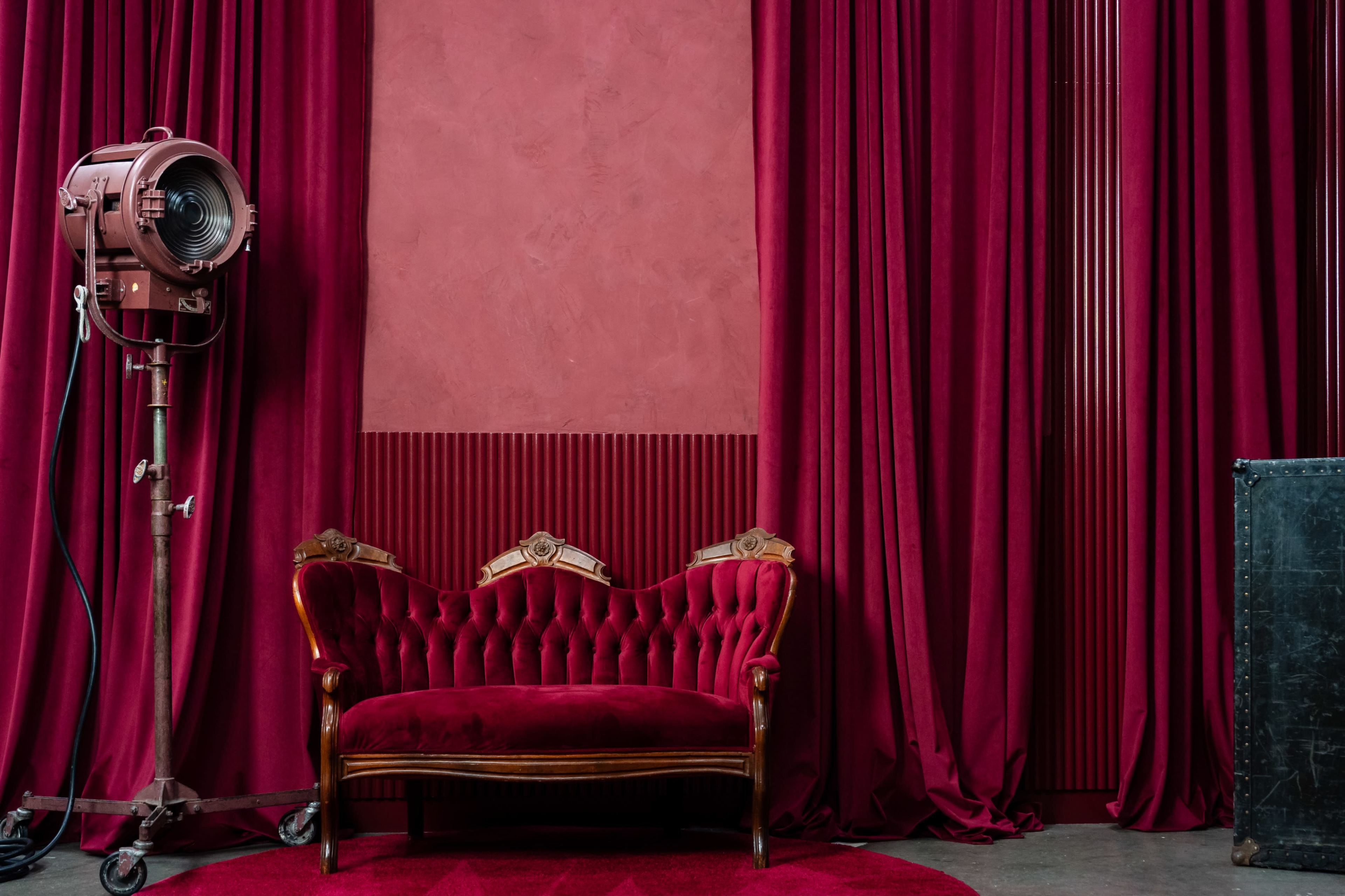 The scene features a red velvet couch in front of matching curtains, with a vintage lighting fixture to the left and a textured wall in the background.