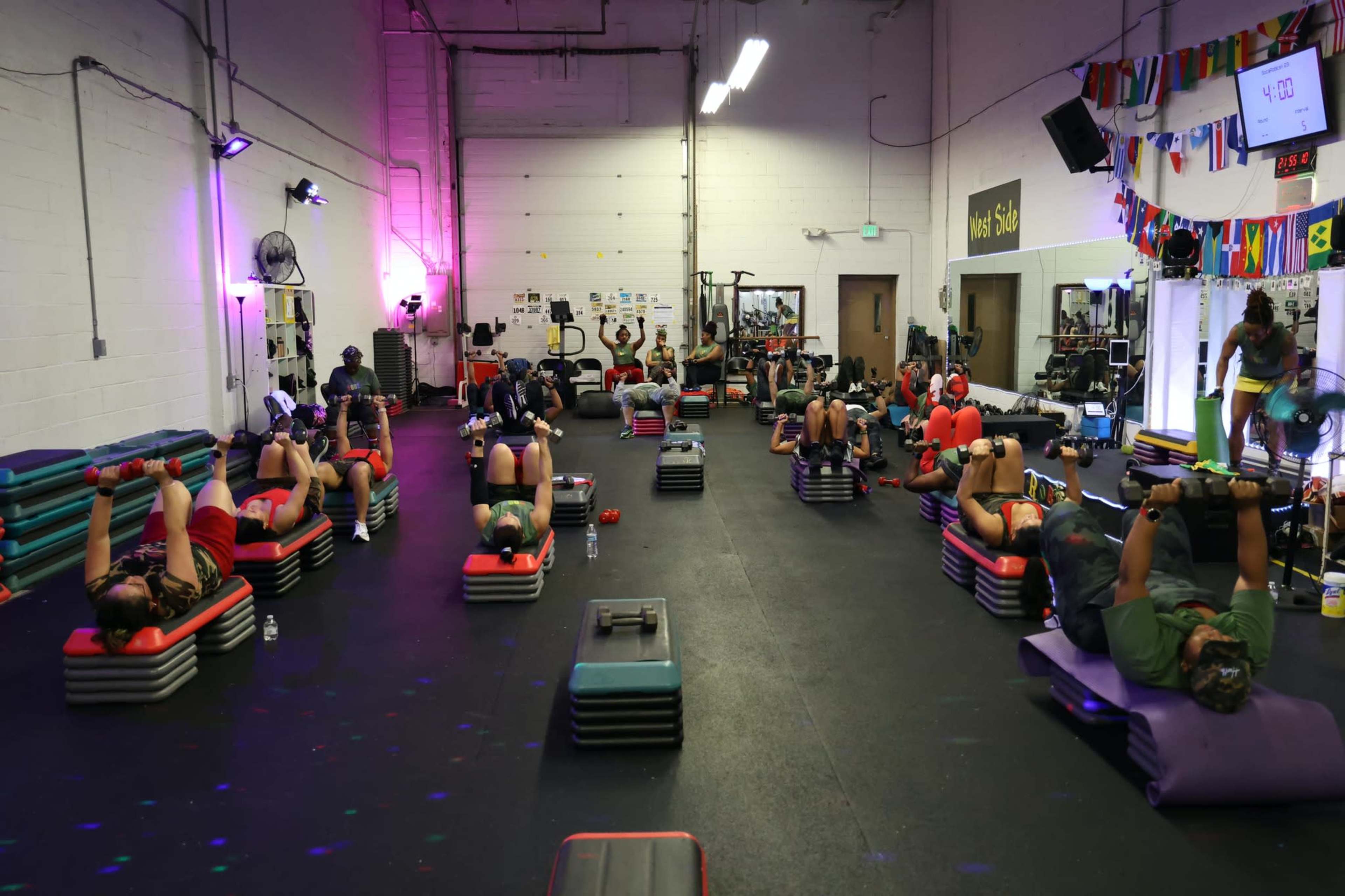 A group of people participates in a fitness class, lying on exercise mats and using weights in a gym setting.