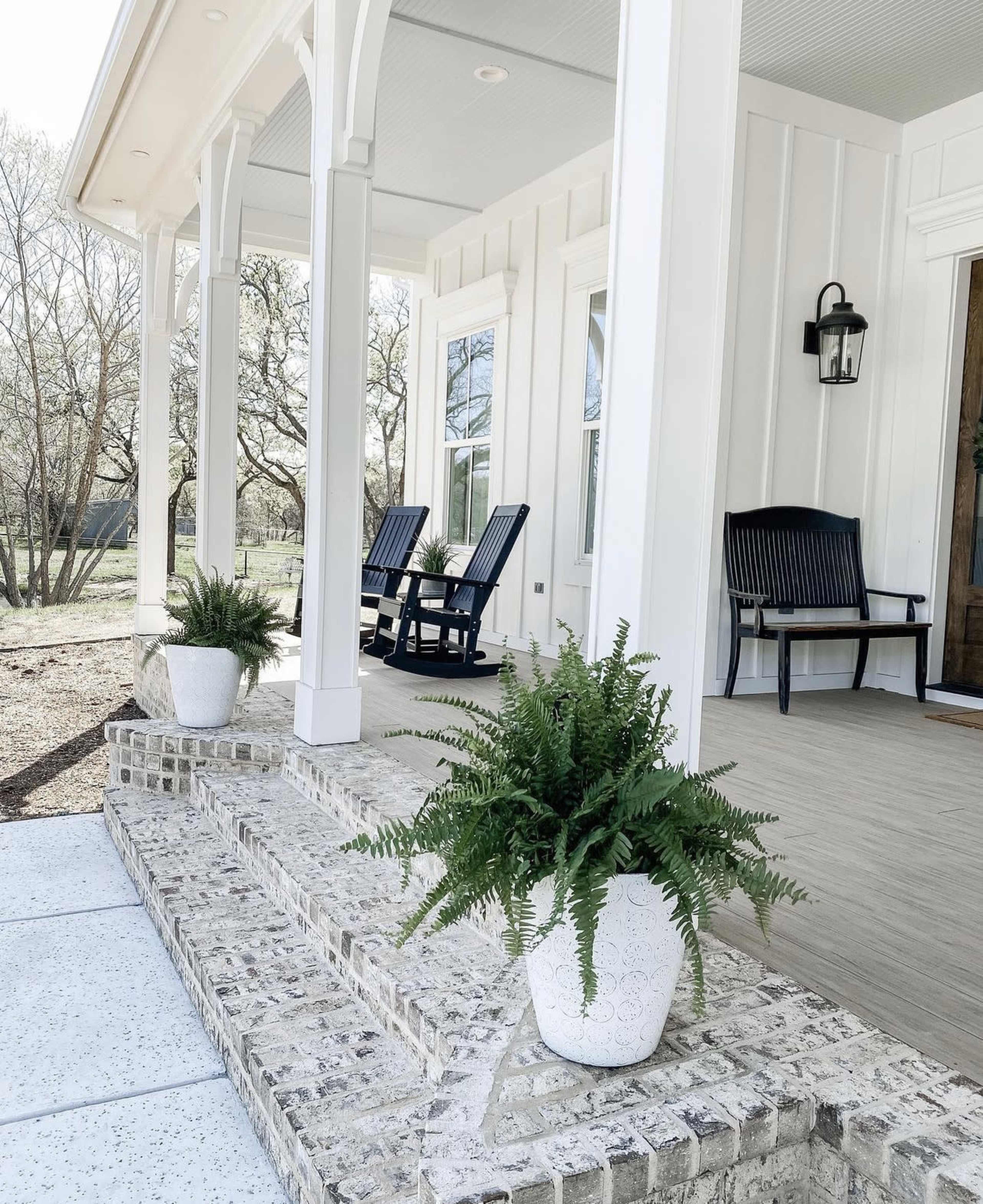 The image shows a home entrance with two rocking chairs on a porch, flanked by potted ferns on brick steps.