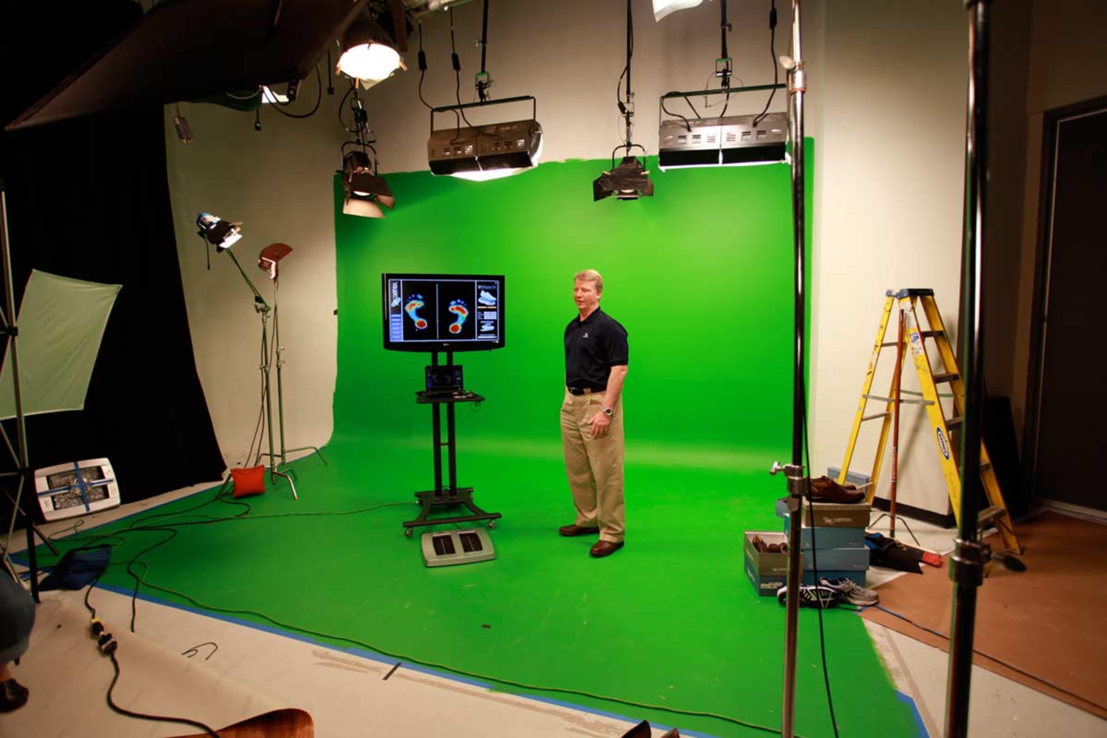 A man stands in front of a green screen, next to a monitor displaying graphics, with studio lights and equipment arranged around the space.