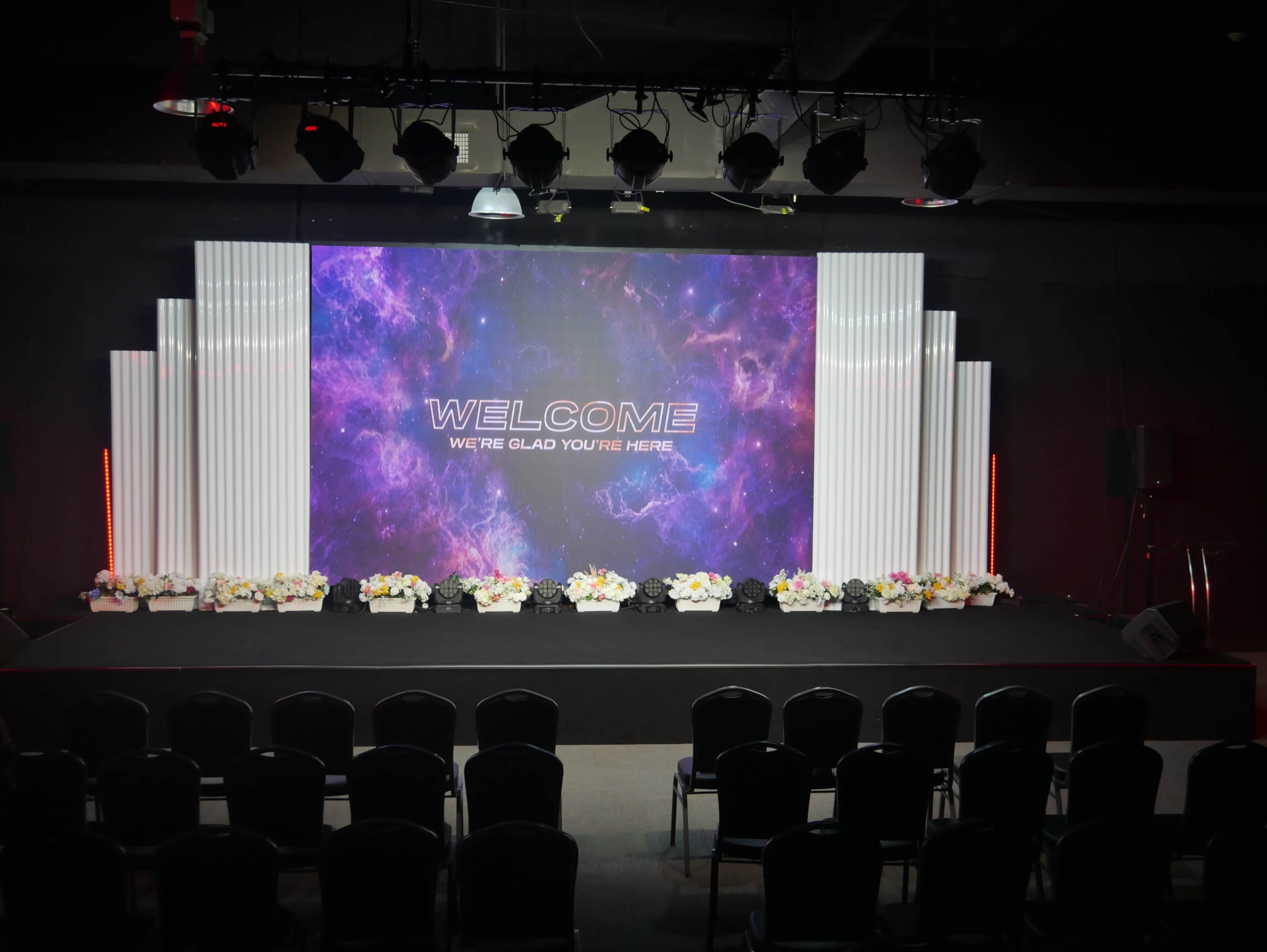 The image shows a stage setup with a large screen displaying "WELCOME" and flower arrangements in front of black chairs positioned for an audience.