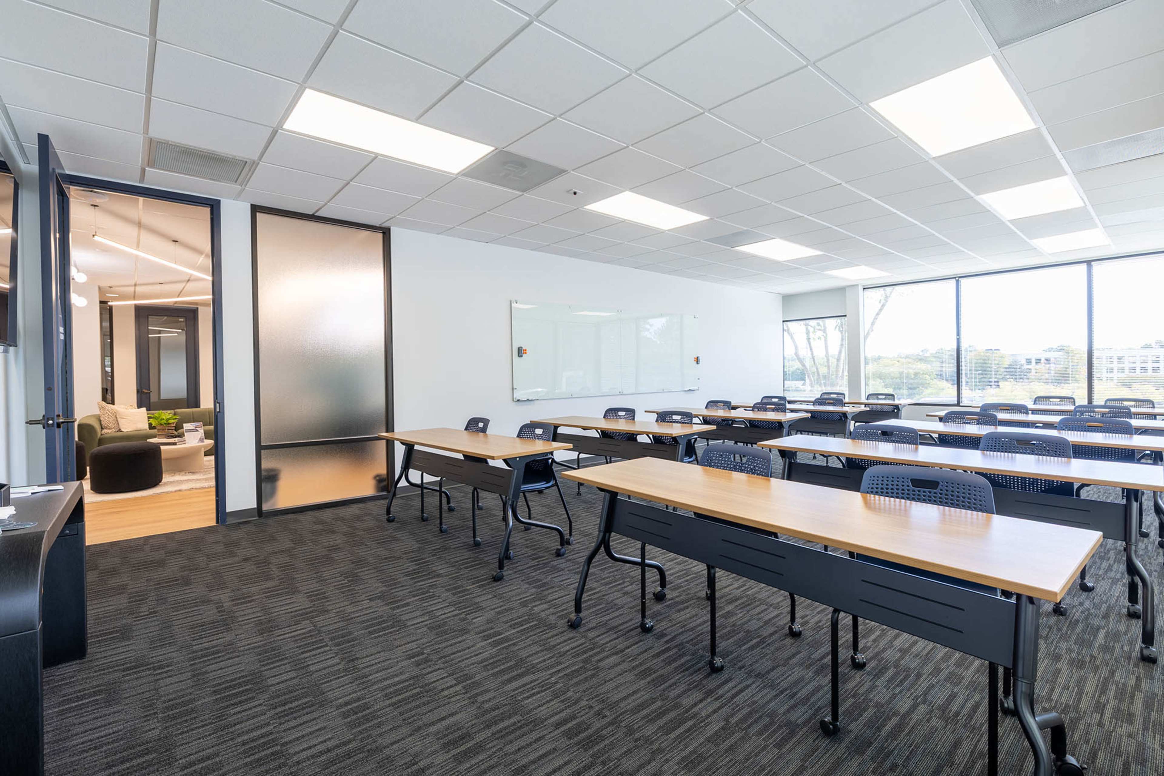 The image shows a bright, modern classroom with rows of desks arranged for a seminar, large windows providing natural light, and a glass-walled office area visible in the background.