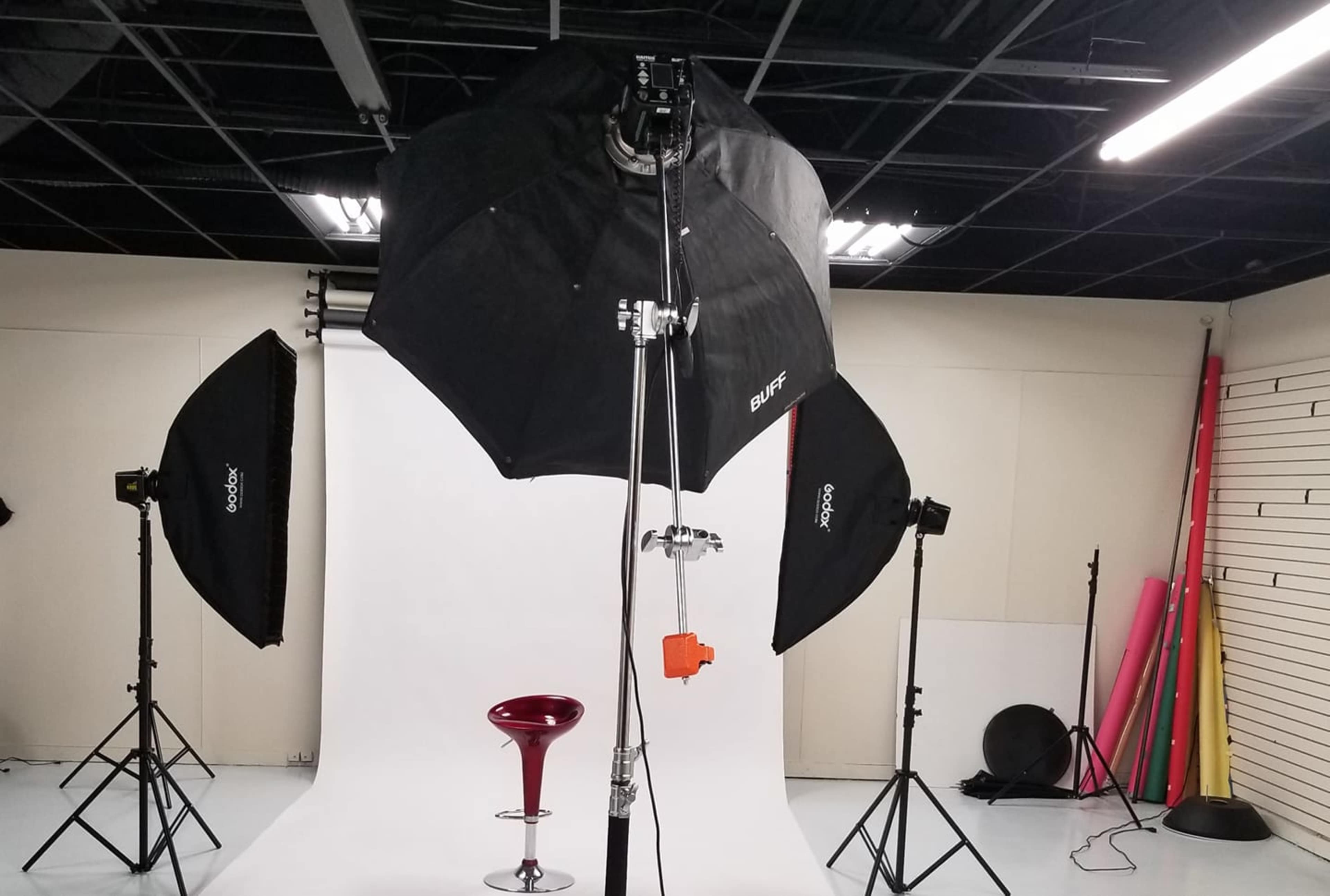 A photography studio is set up with an empty red stool in front of a white backdrop, illuminated by three softbox lights.