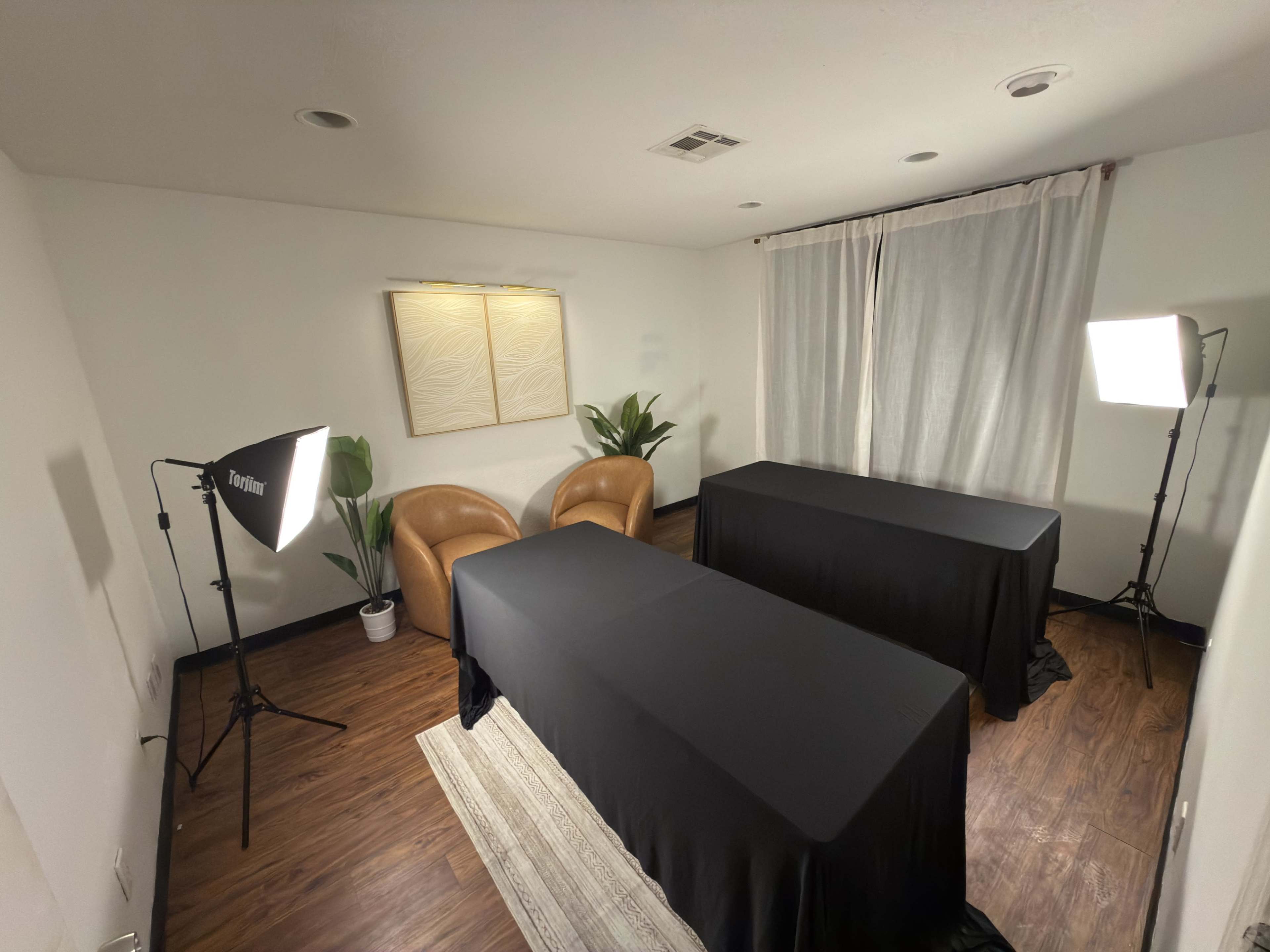 The image shows a well-lit room with two tables covered in black tablecloths, flanked by two brown chairs and potted plants, with overhead lighting and a curtain window in the background.