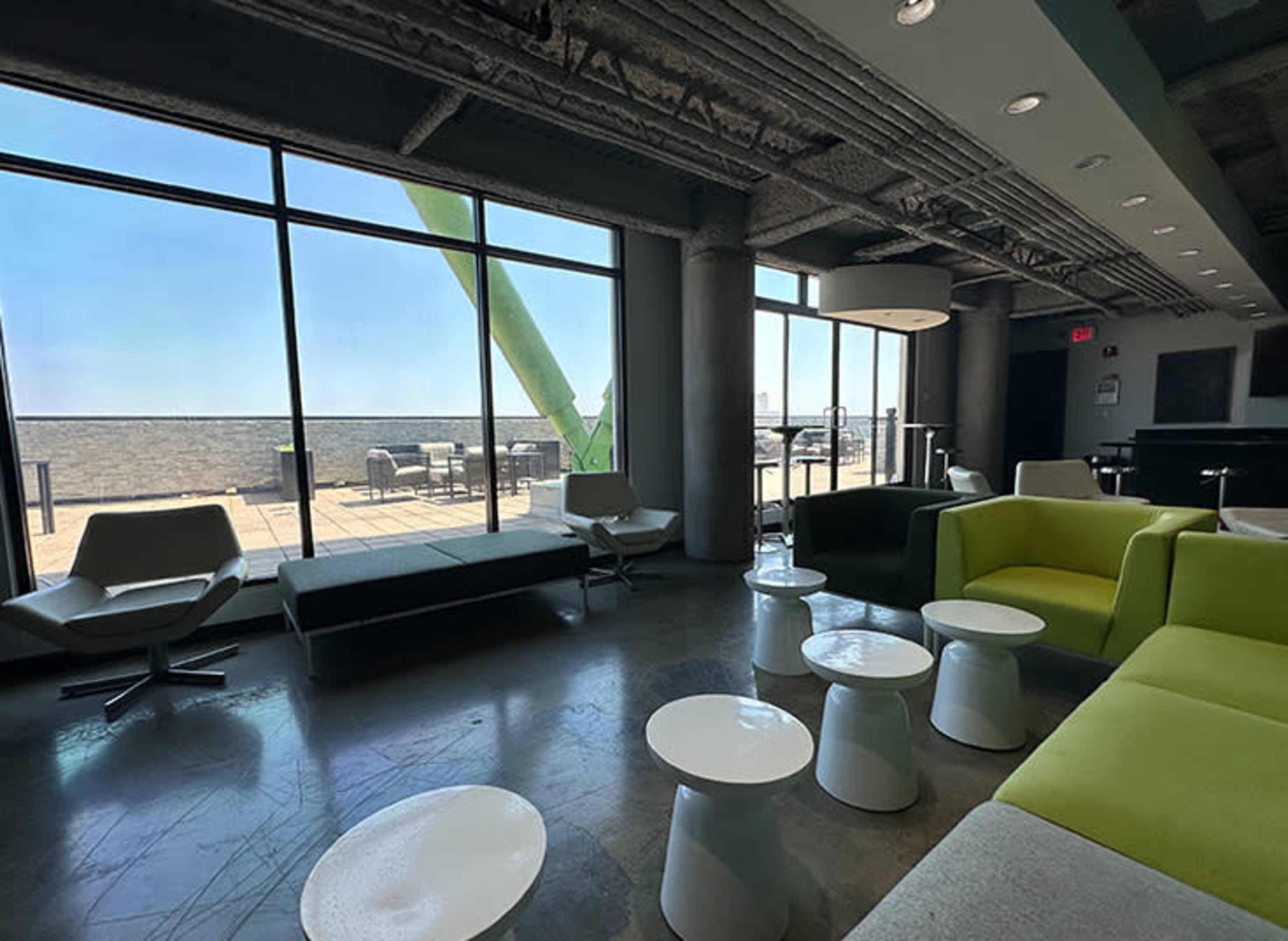 The image depicts a modern lounge area with large windows showcasing a patio, featuring a mix of seating options in light colors and small white tables.