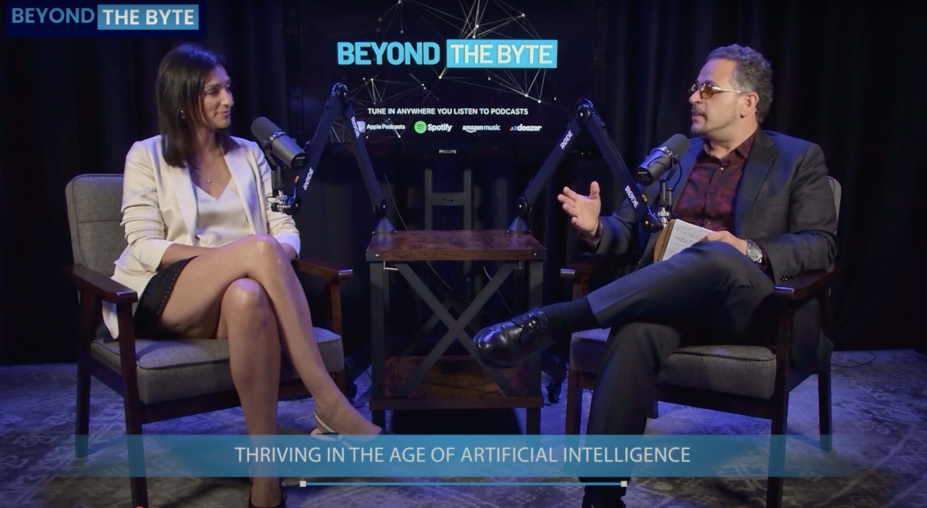 A male and female speaker are seated in chairs, engaged in a discussion at a podcast setup featuring microphones and a backdrop that reads "BEYOND THE BYTE."