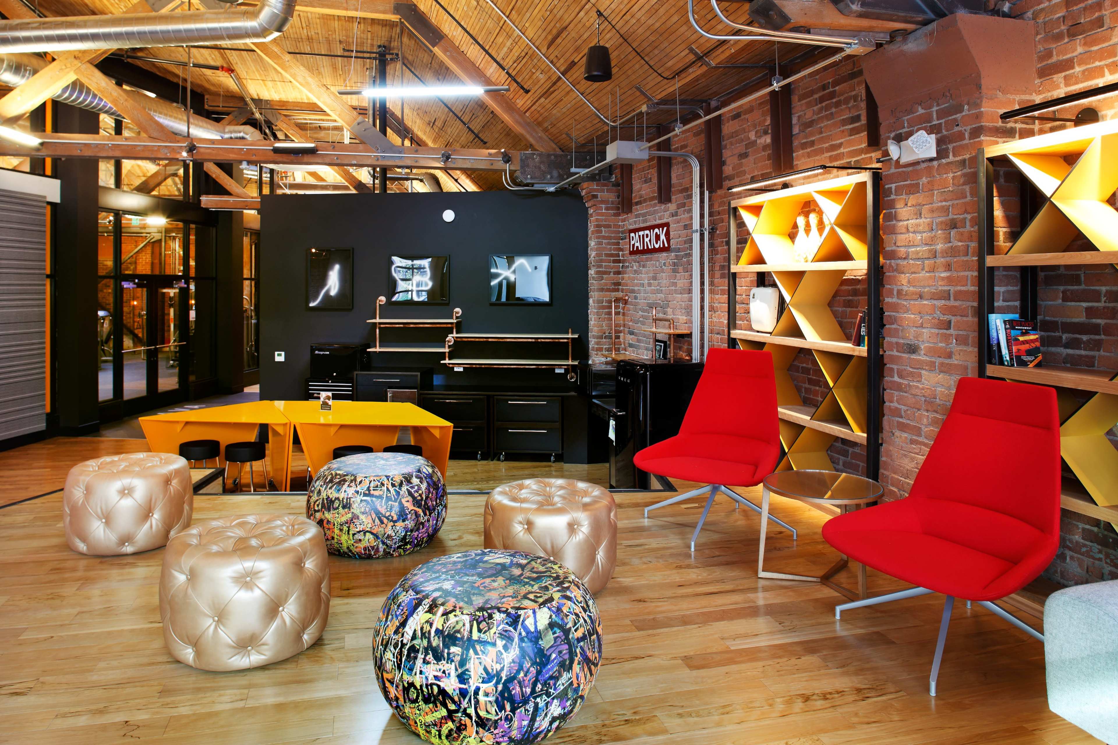A modern interior space with wooden flooring, exposed brick walls, a black kitchenette, colorful seating options, and shelves displaying various items.