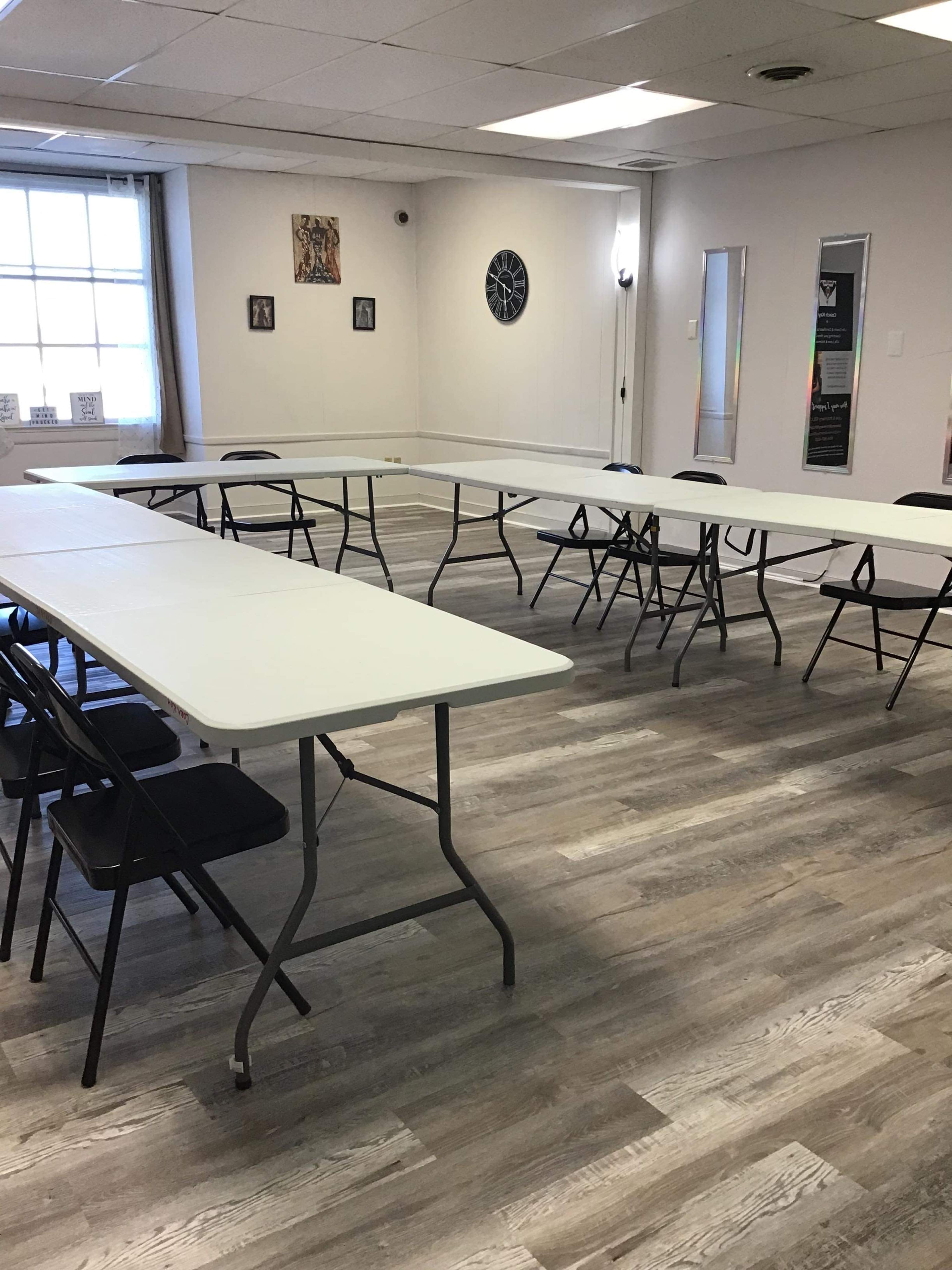 A room set up with several folding tables arranged in a U-shape, surrounded by black chairs, with windows and wall decorations in the background.