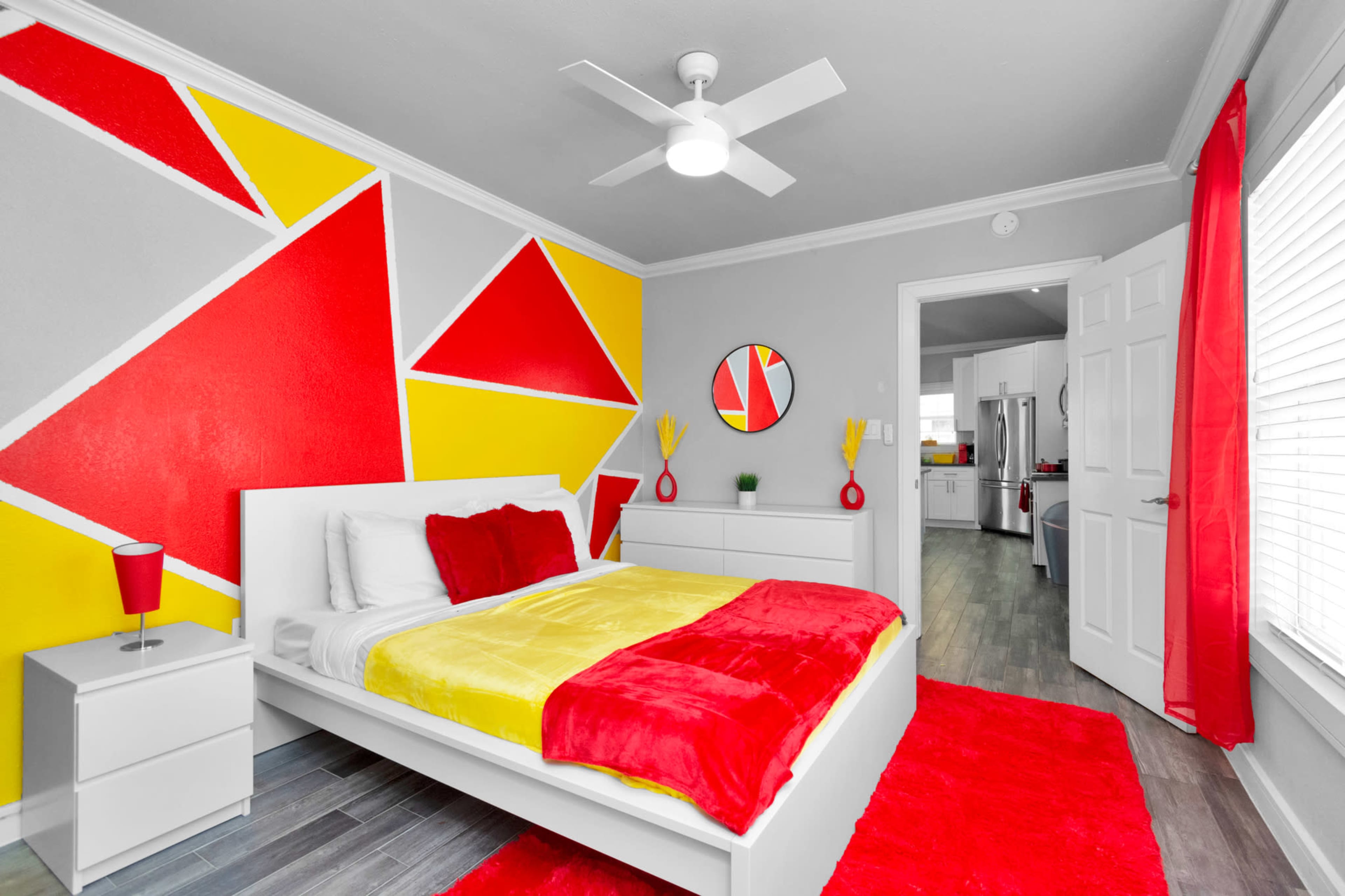 A modern bedroom featuring a white bed with red and yellow bedding, geometric wall art in red and yellow, a white nightstand, and red curtains.
