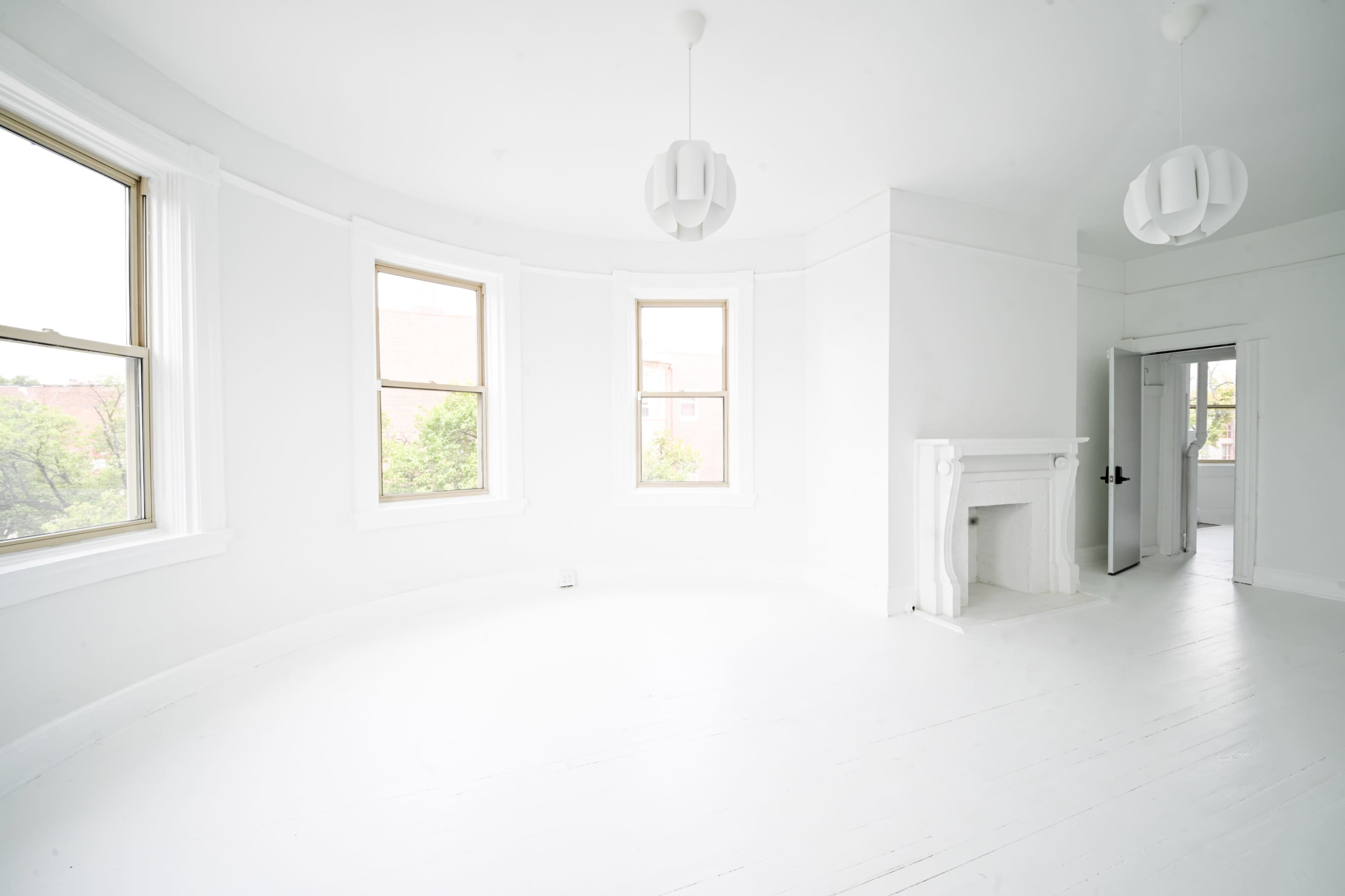 A bright, empty room with white walls and flooring features three windows and two pendant light fixtures.