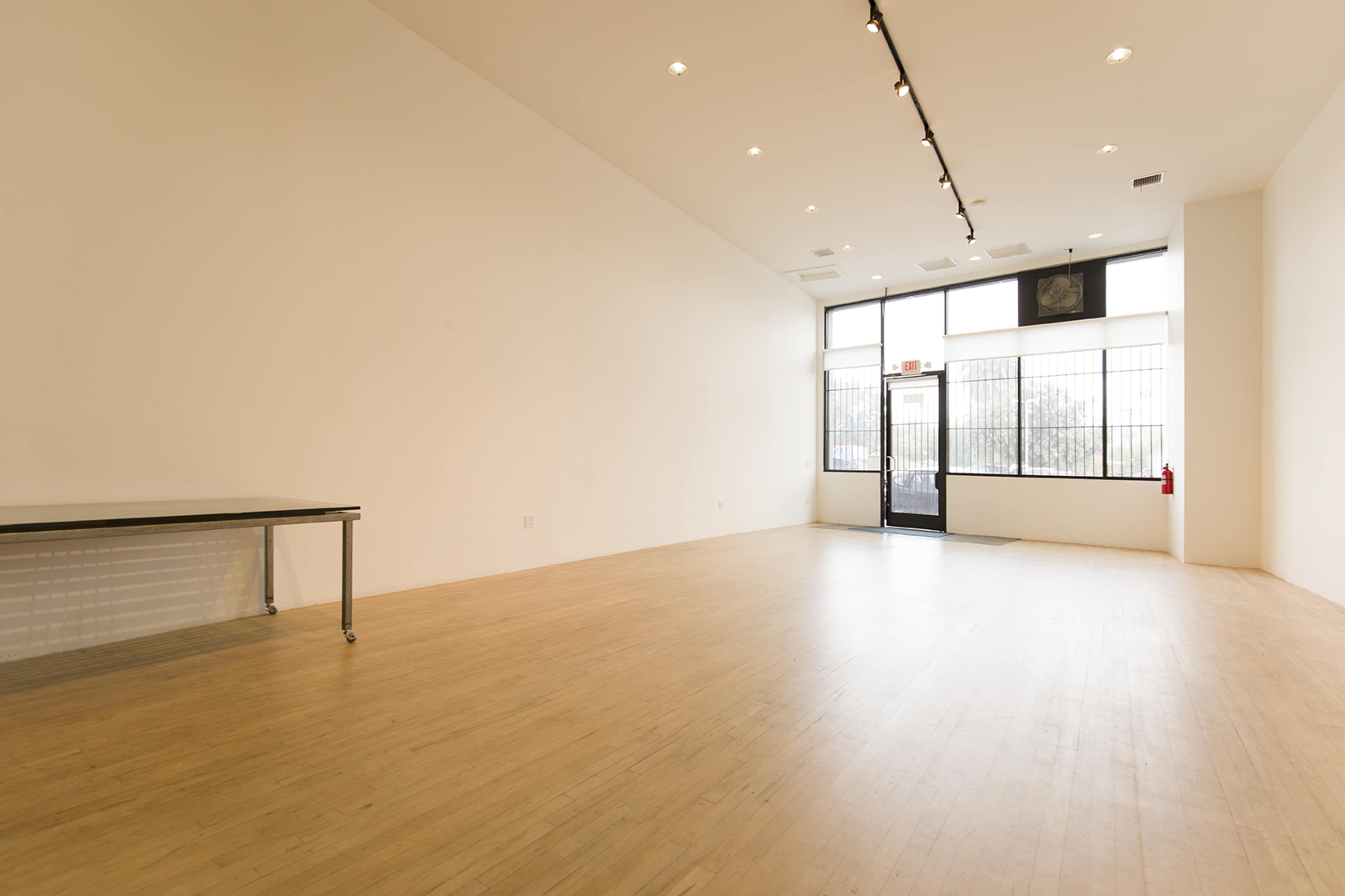 The image shows a spacious, empty room with wooden flooring, a table on one side, and large windows providing natural light.