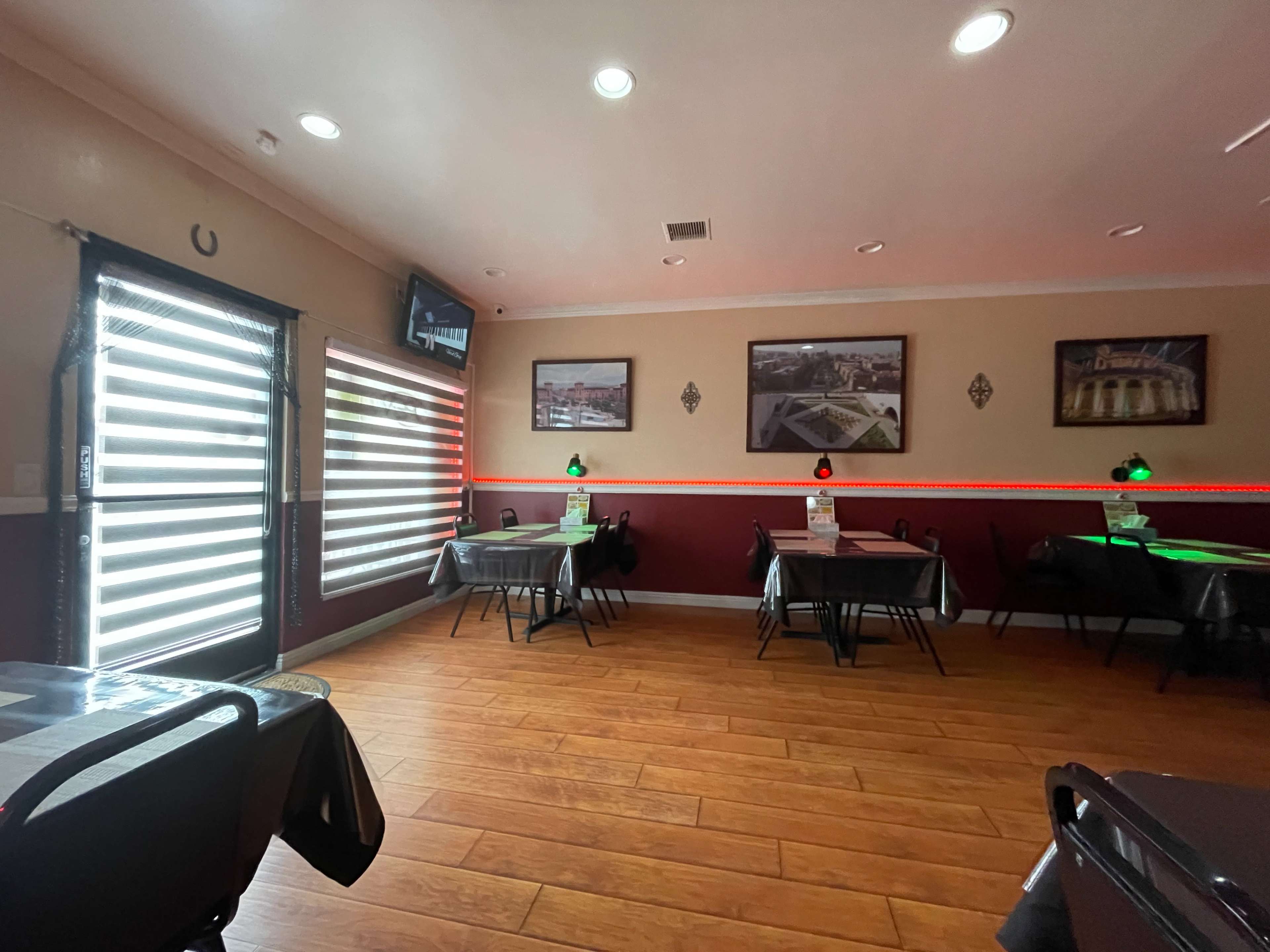 The image shows the interior of a restaurant with several tables, featuring window blinds, wall decorations, and ambient lighting.
