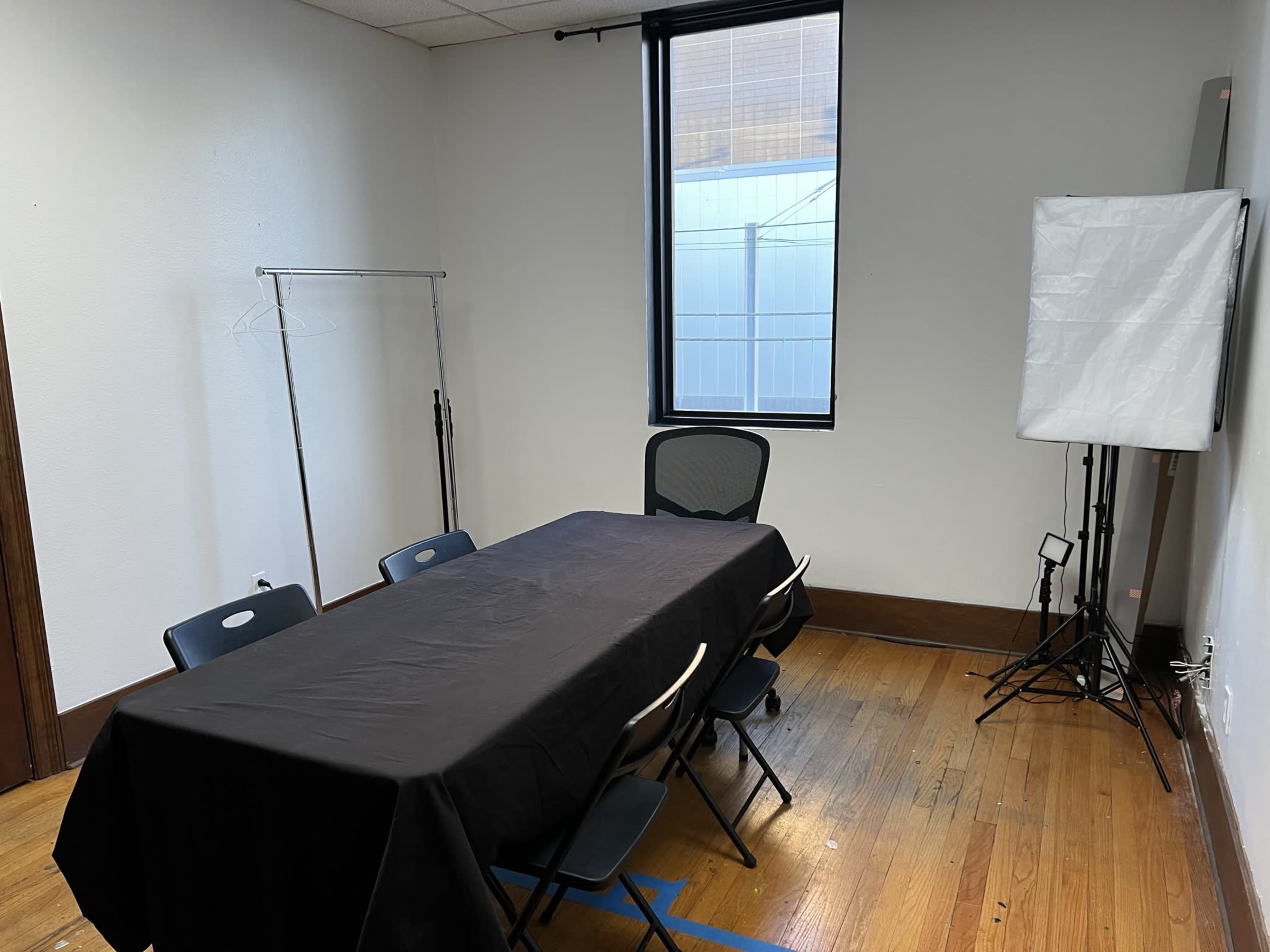 A small meeting room features a large table covered with a black cloth, surrounded by chairs, and illuminated by a light setup near a window.