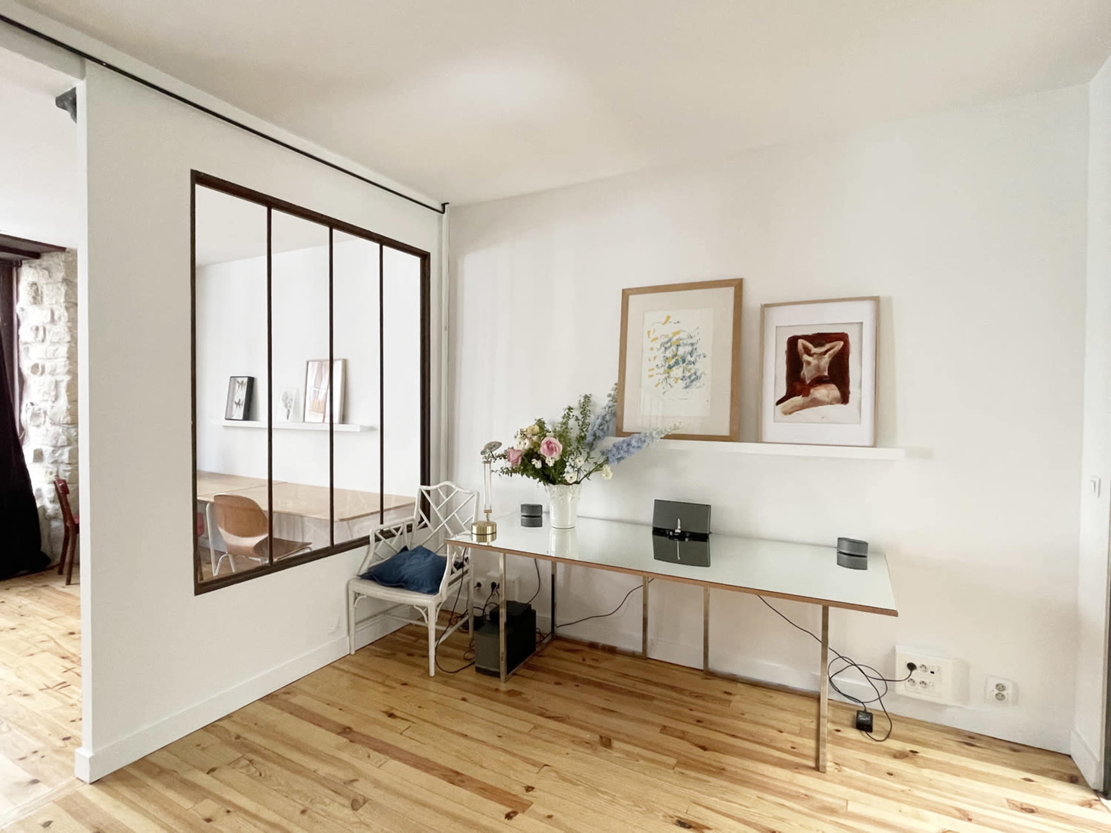A modern workspace with a glass desk, decorative artwork on the wall, and a floral arrangement placed on the desk, adjacent to a partition with windows.