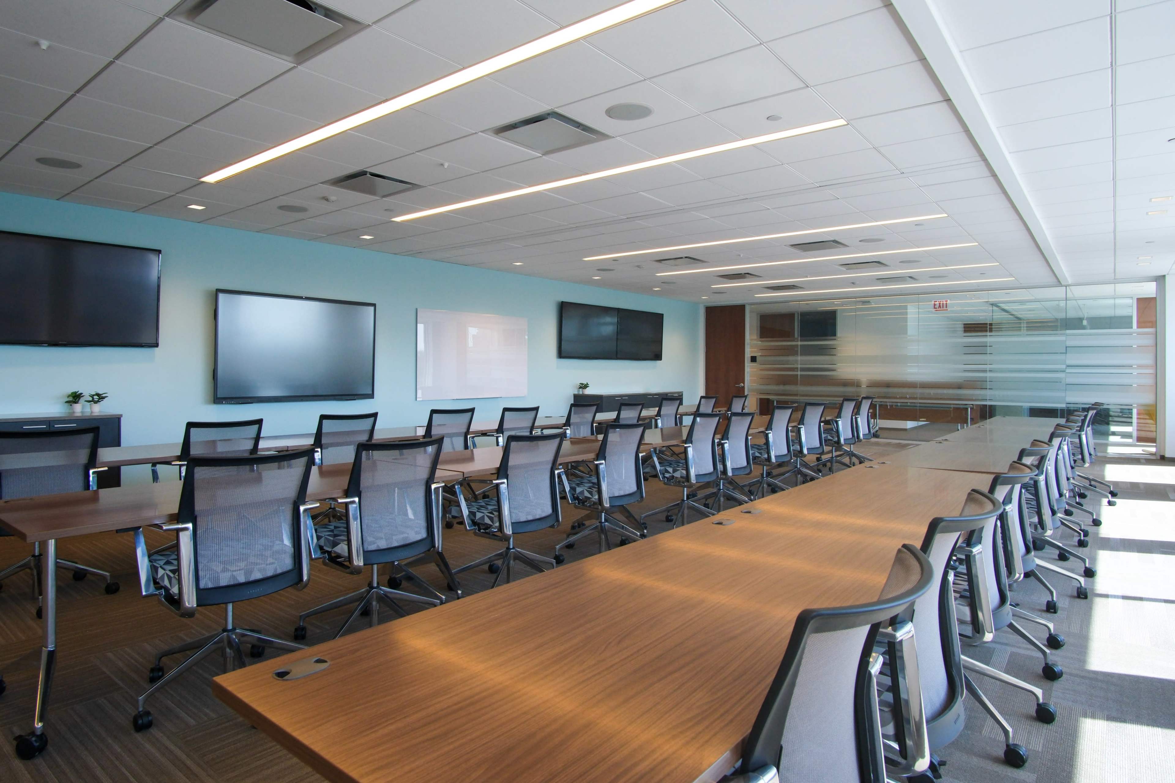 The image shows a modern conference room with a long table, multiple chairs, and several screens mounted on the walls.