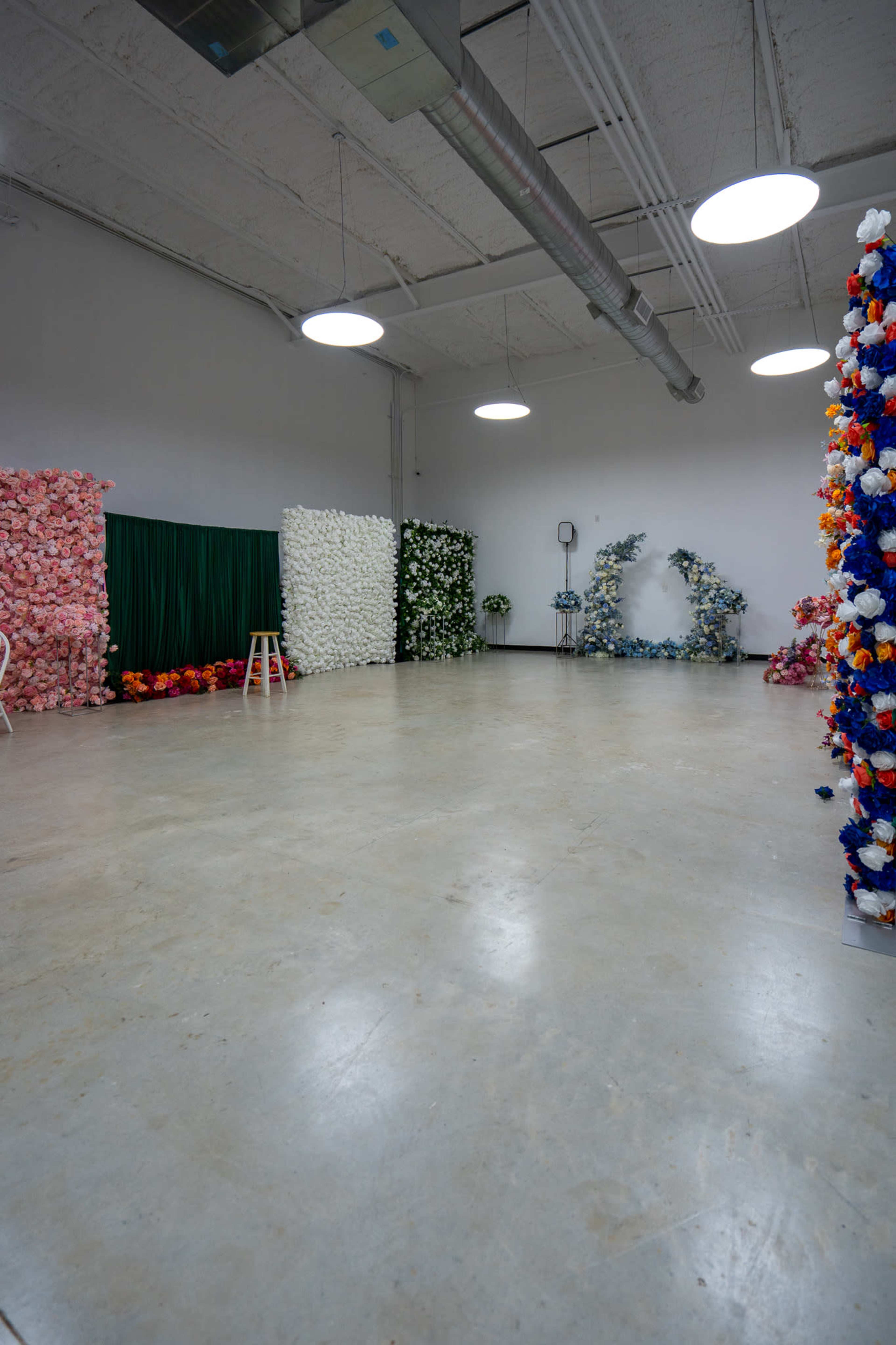 The image shows a spacious room with white walls, a polished concrete floor, and four colorful backdrops made of artificial flowers.