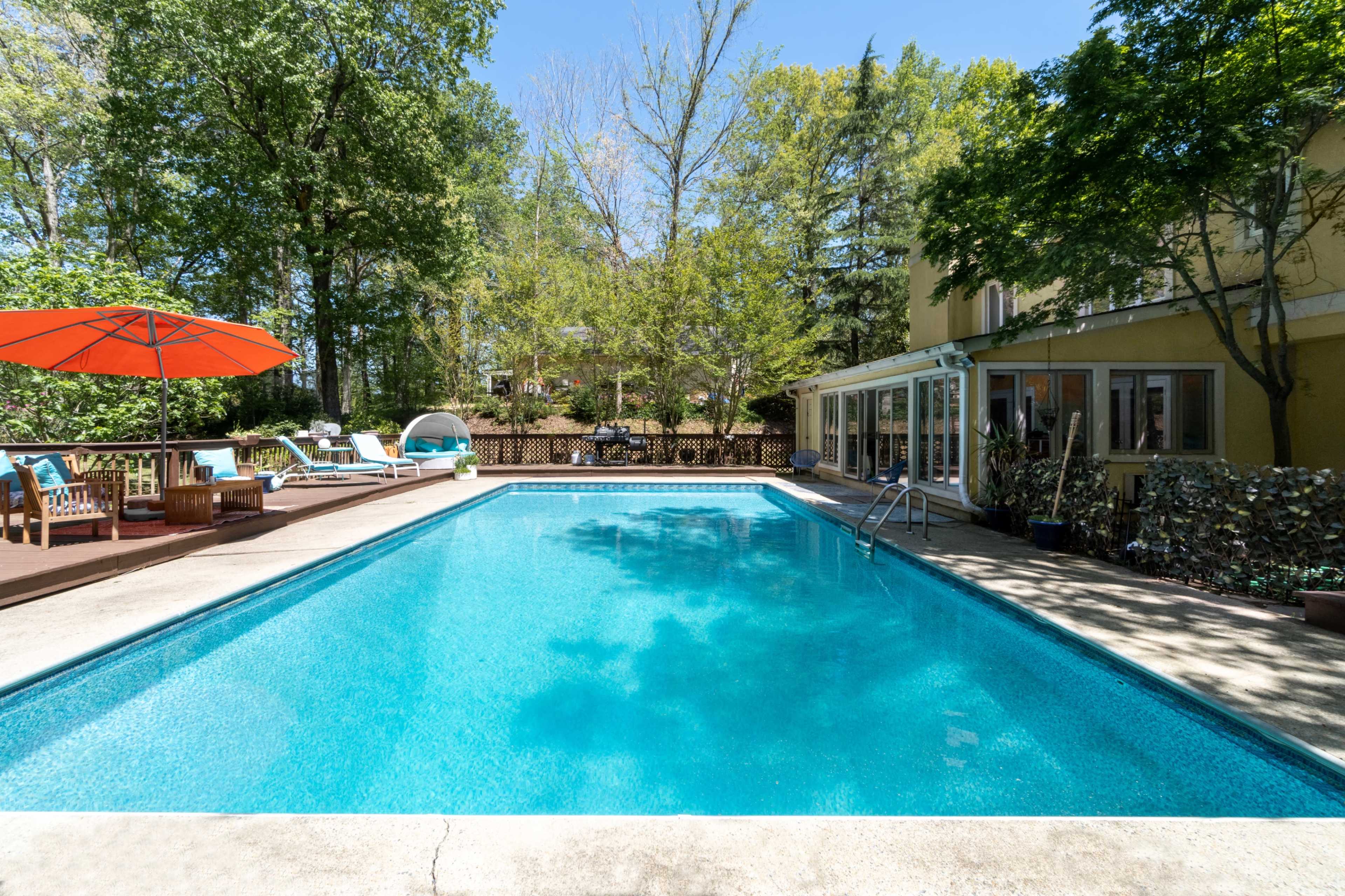 A clear swimming pool is surrounded by lounge chairs and an umbrella, with a house and trees visible in the background.