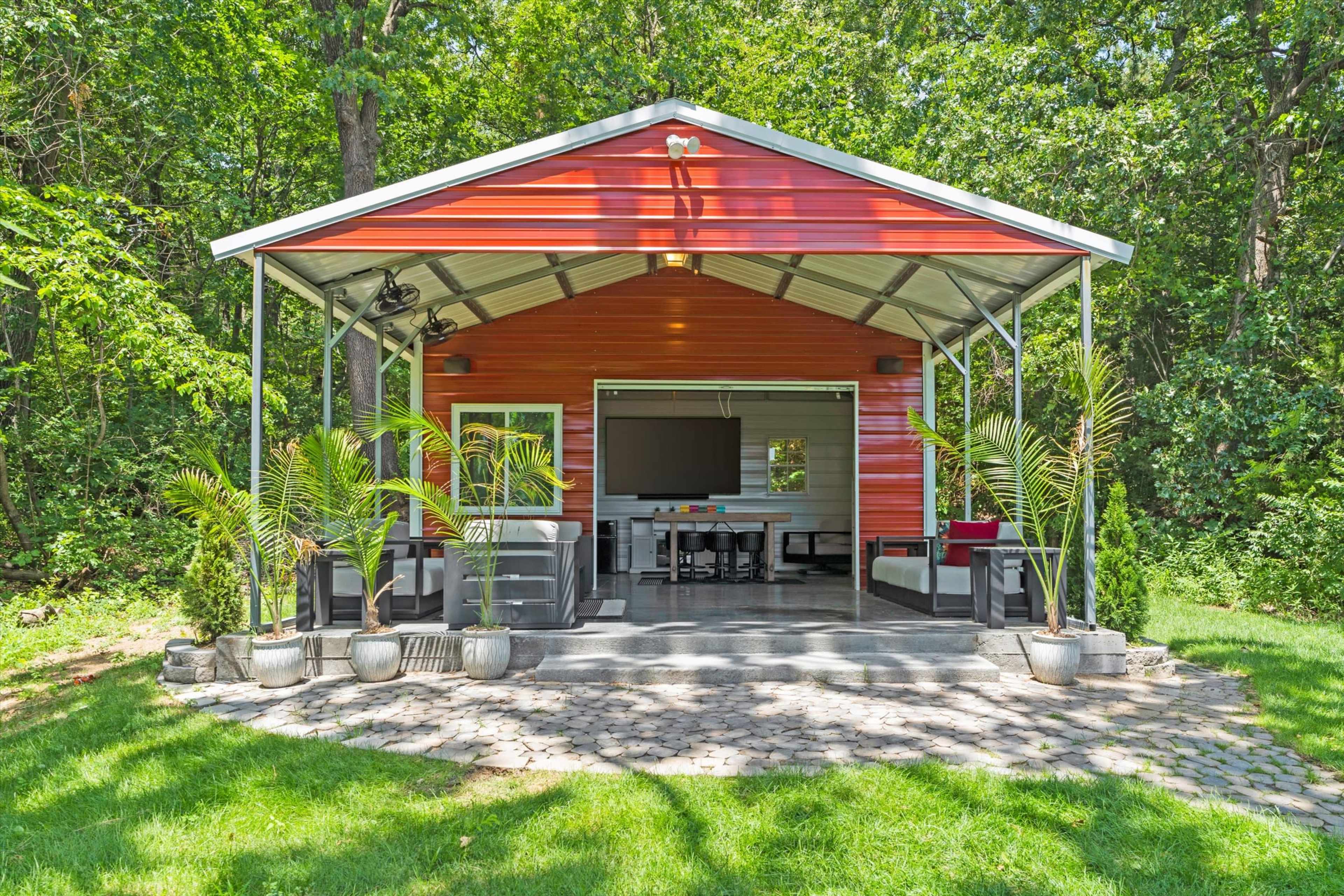 A red and gray outdoor pavilion, featuring a seating area, a television, and surrounded by greenery and stone pavement.