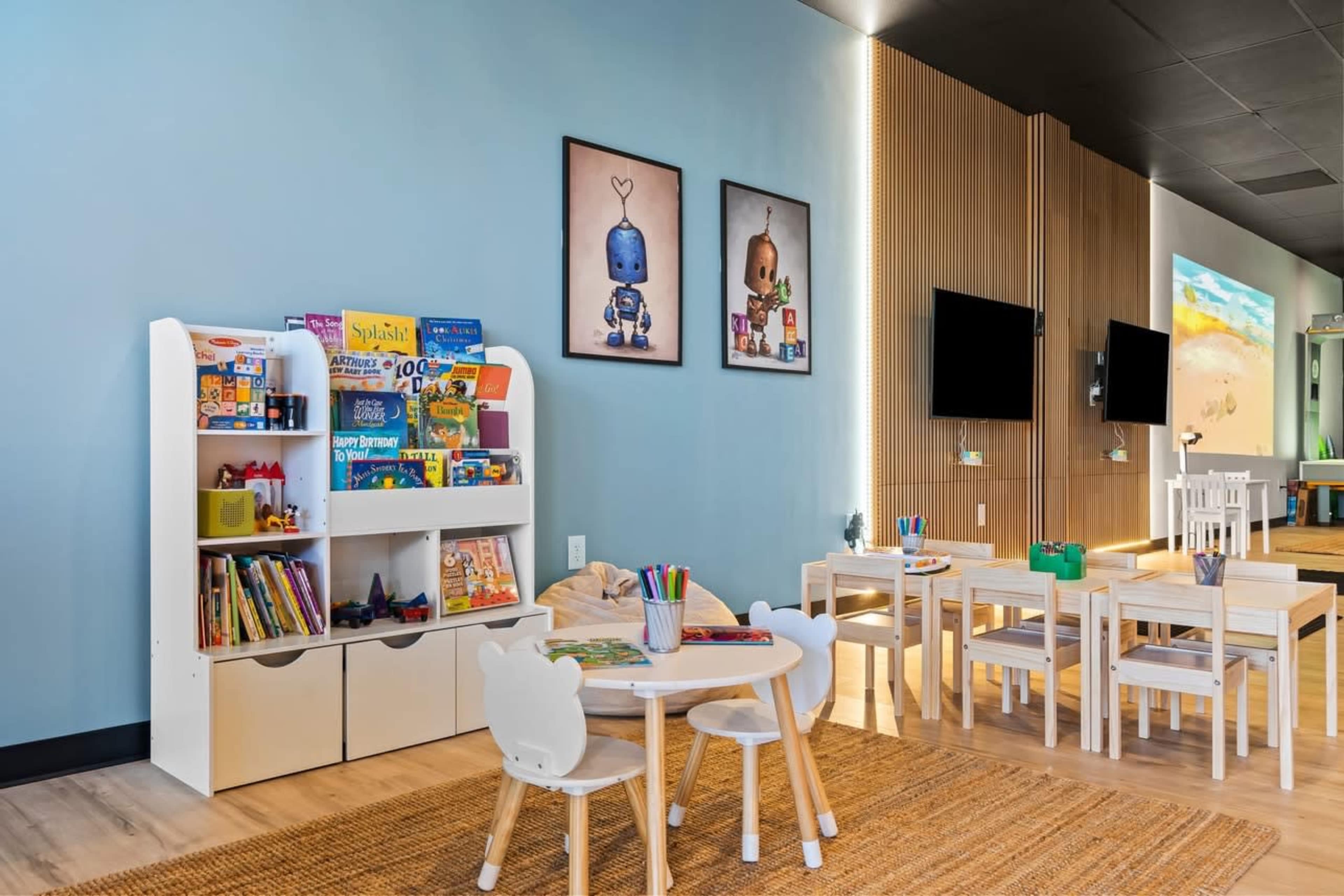The image shows a brightly lit children's playroom with a bookshelf filled with toys and books, and several small tables and chairs arranged on a wooden floor.