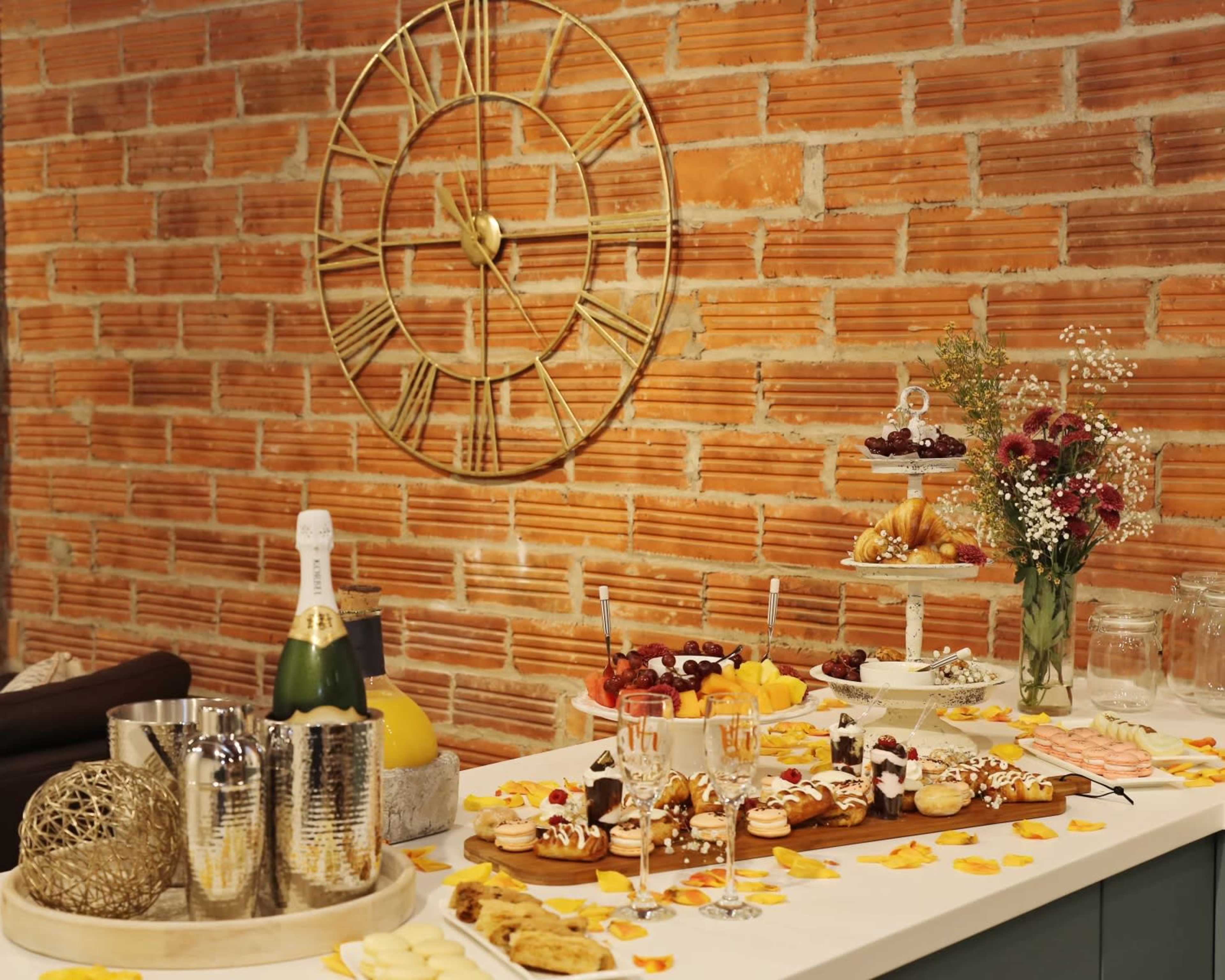 A buffet table is set against a brick wall, featuring various desserts, fruits, and a bottle of champagne alongside decorative items.