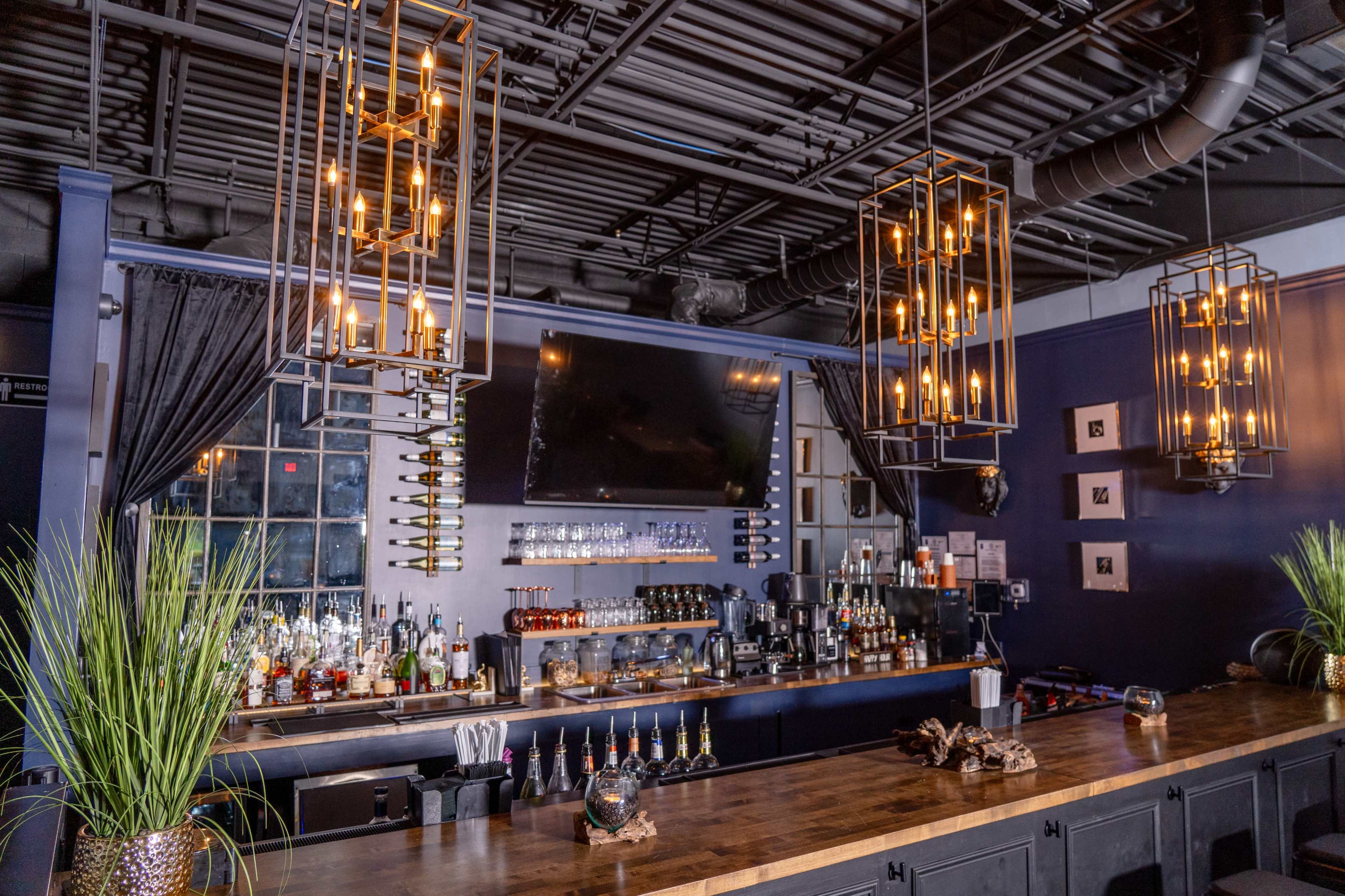 A modern bar with a long wooden counter, overhead pendant lights, a selection of bottles on the shelves, and a large TV screen mounted on the wall behind it.