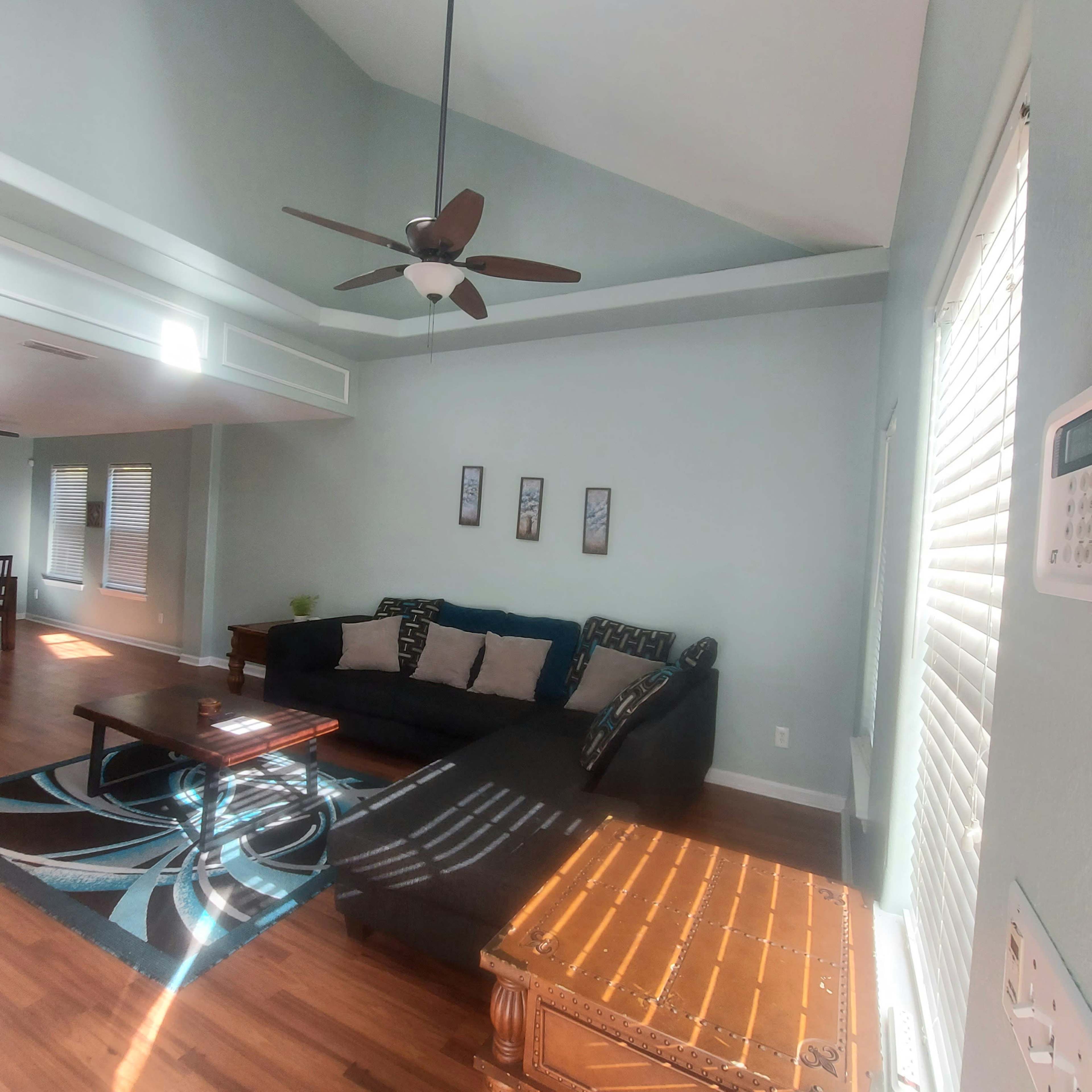 A living room with a ceiling fan, a black sectional sofa, a coffee table, and large windows letting in natural light.