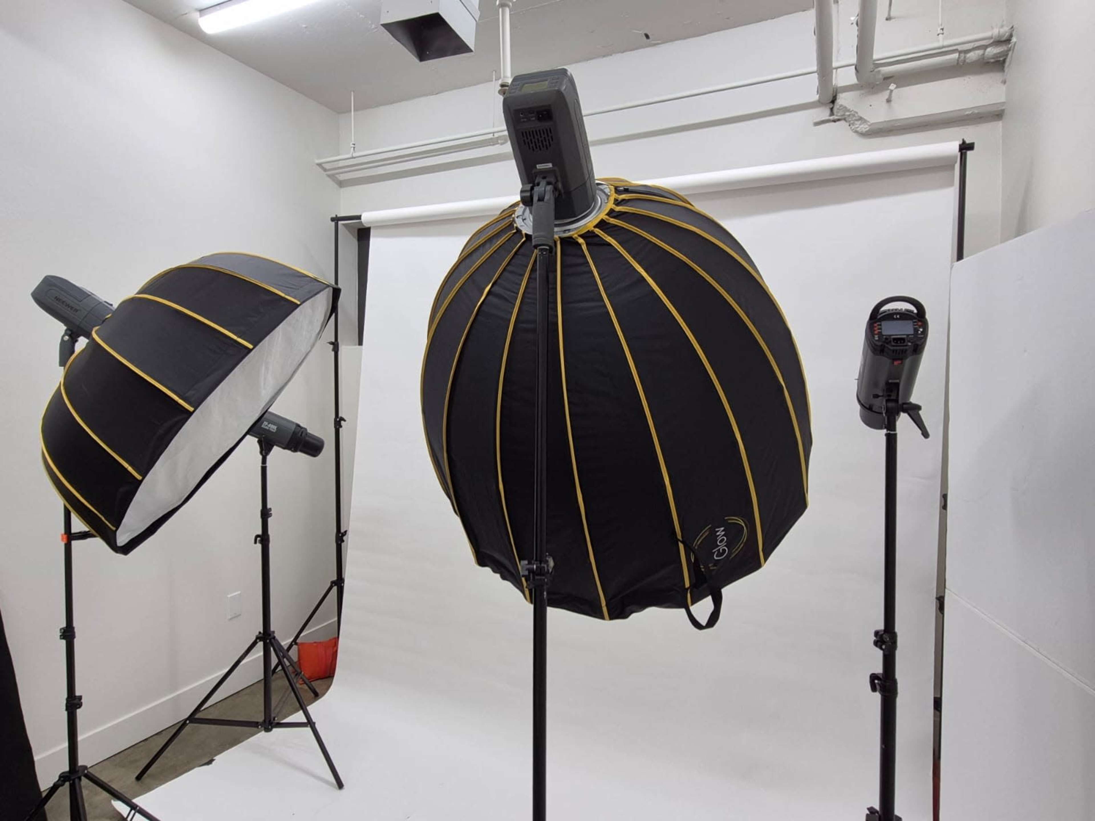 The image shows a photography studio setup with two large softbox lights and a camera on a tripod, all positioned around a white backdrop.