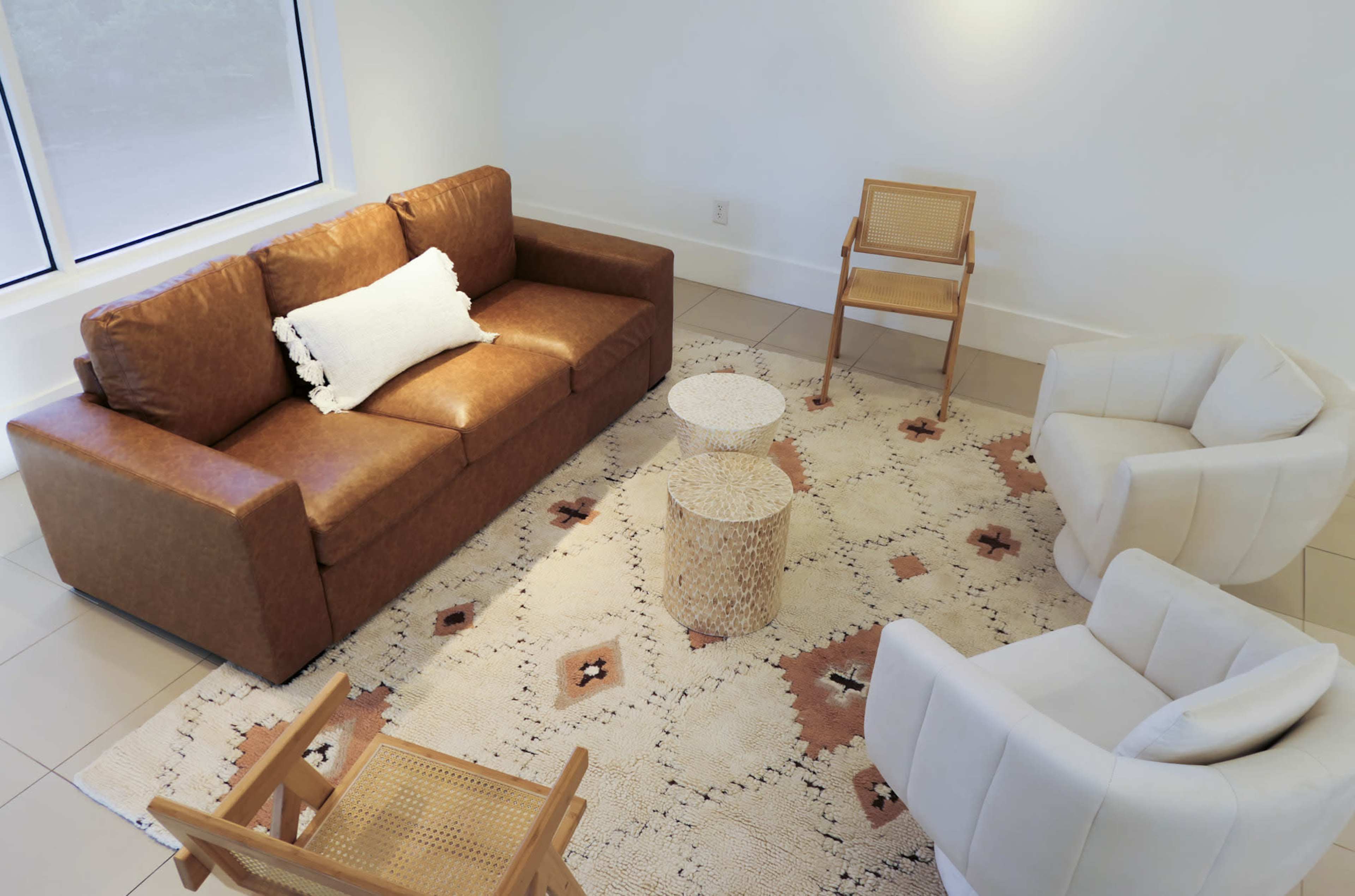 A cozy seating area features a brown leather sofa, two white armchairs, a textured side table, and a patterned rug.