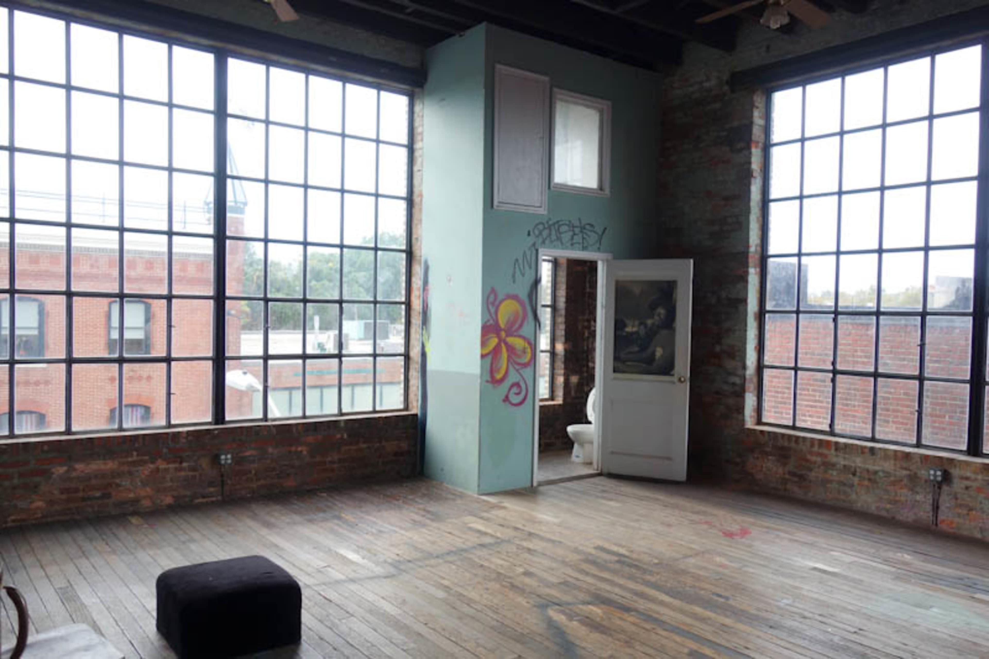 A spacious, industrial-style room with large windows, exposed brick walls, and a door leading to a small bathroom.