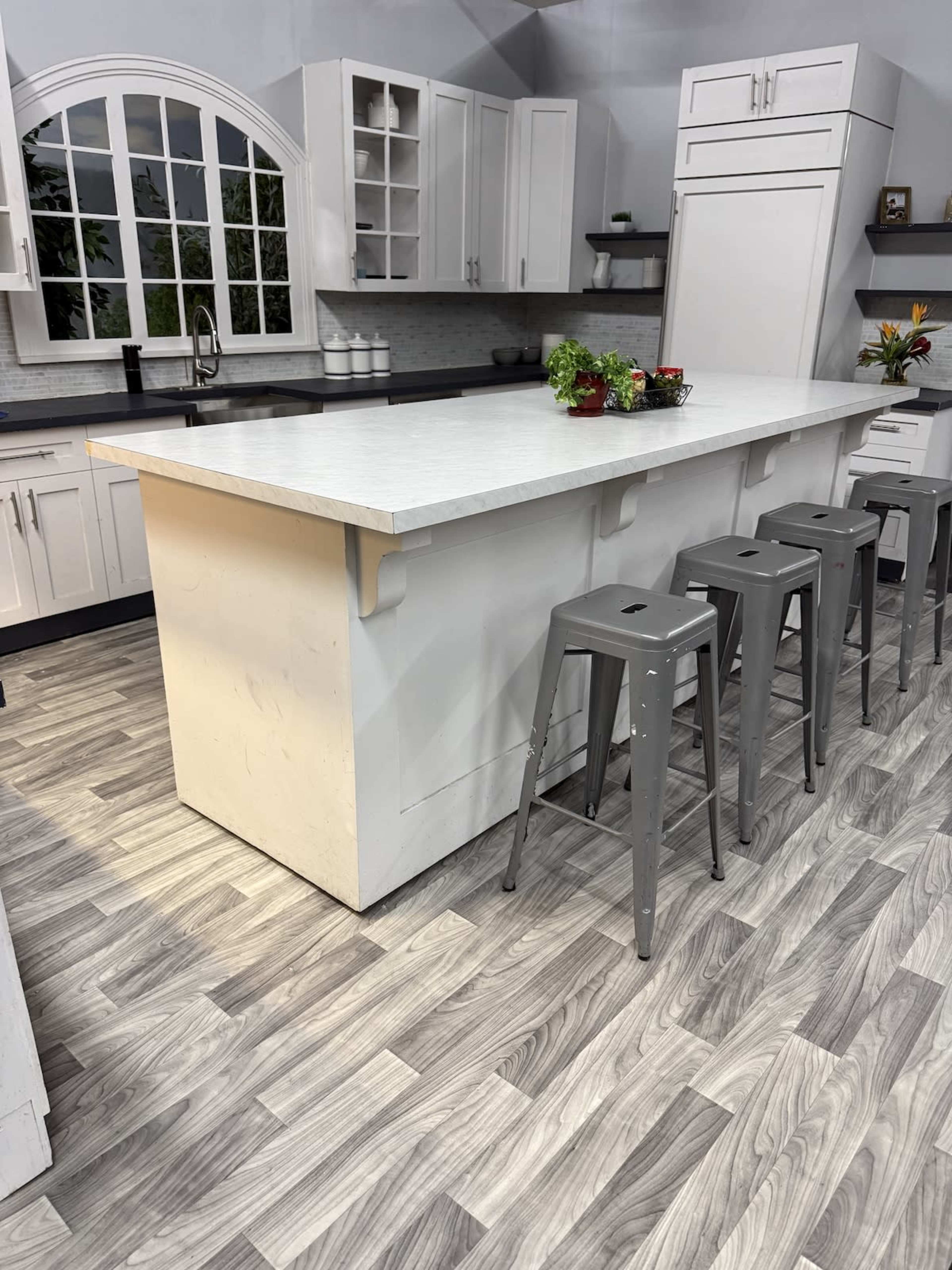 The image shows a modern kitchen with a large island and metal stools, featuring a sleek countertop and light-colored cabinetry.