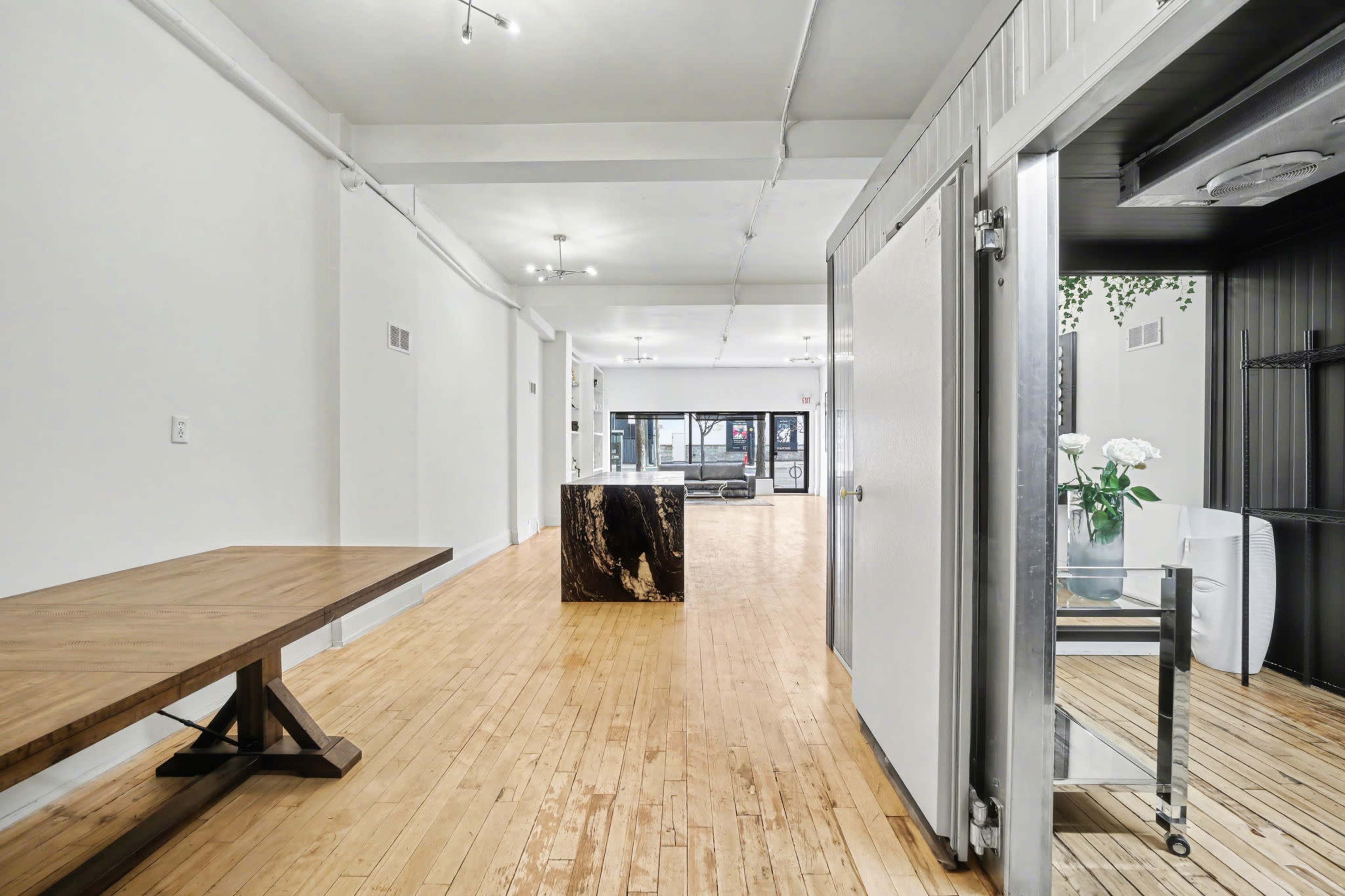 The image shows an open, well-lit interior space featuring a wooden table, a decorative block, and a view into a lounge area.