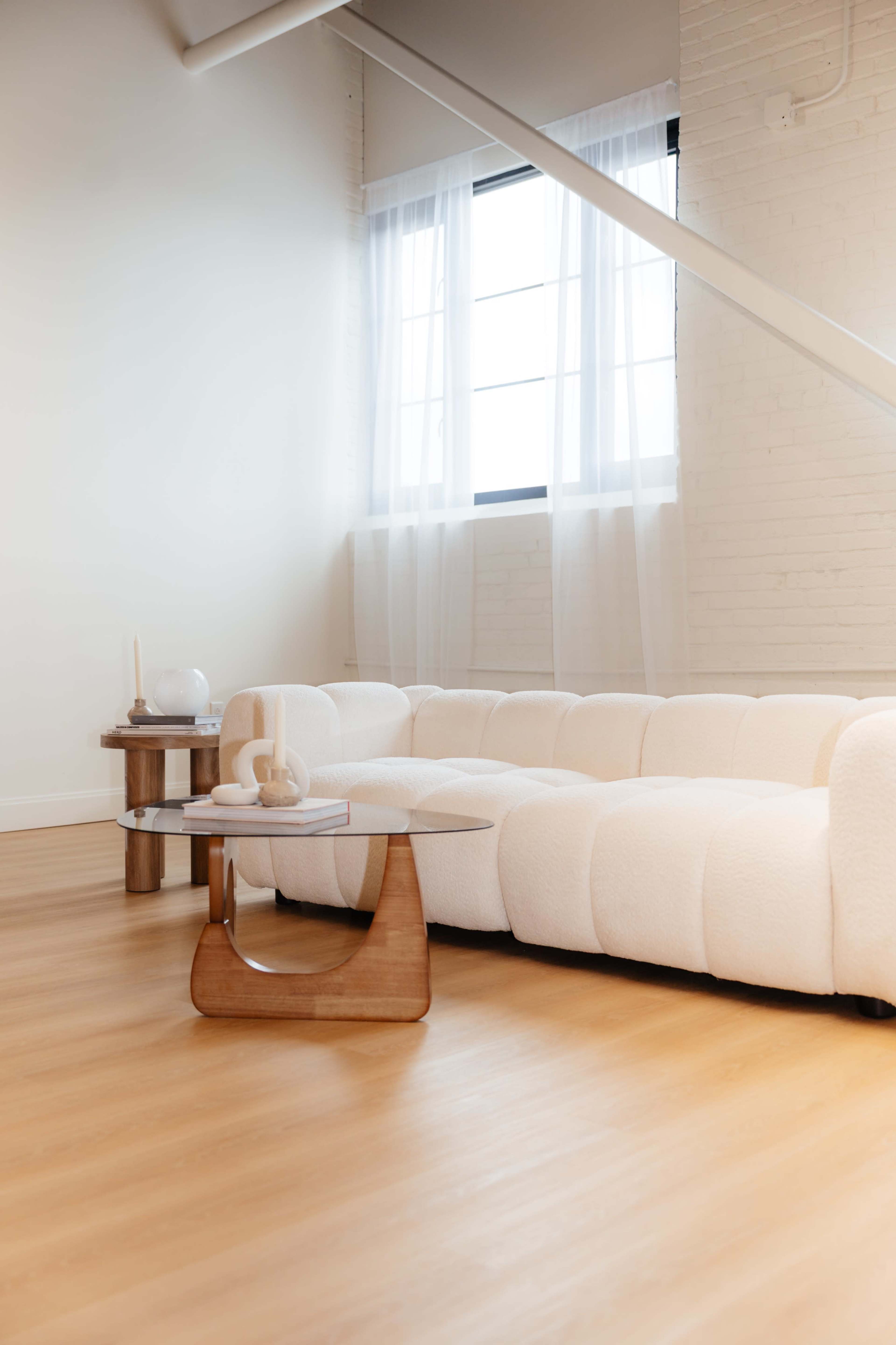 The image shows a modern living room featuring a white, plush sofa, a small wooden coffee table, and large windows with sheer curtains.