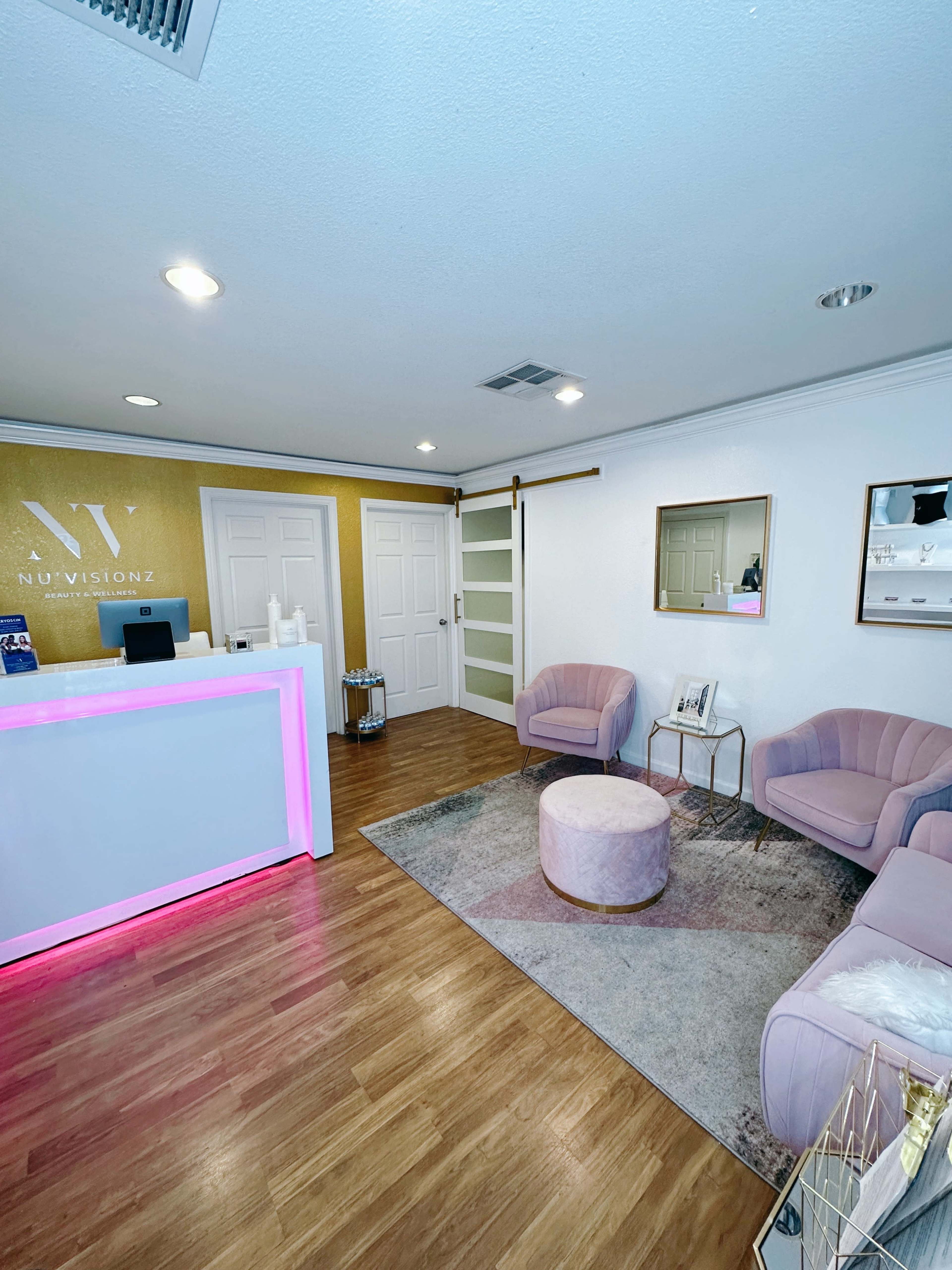 A modern reception area with a white front desk illuminated in pink, two pink chairs, a round ottoman, and a decorative rug.